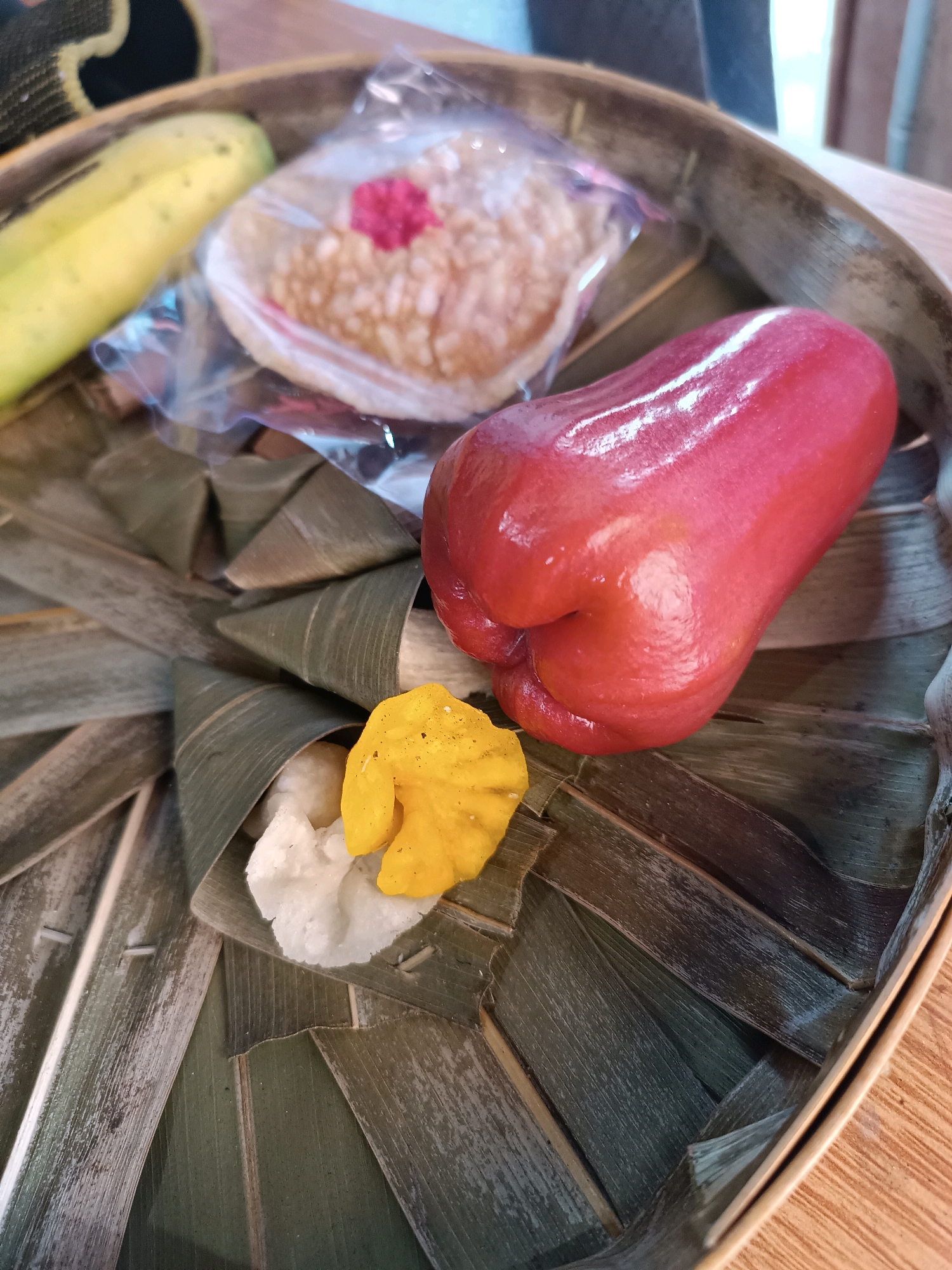 In a woven bamboo basket lined with banana leaves, a tray of food is presented. A bright red rose apple, a yellow flower petal, a piece of white rice, and a round snack are arranged on the leaves. To the left, a banana rests near the edge of the basket. The basket is placed on a wooden surface. This image was taken in Tabanan, Indonesia, likely within a local market or a food stall.