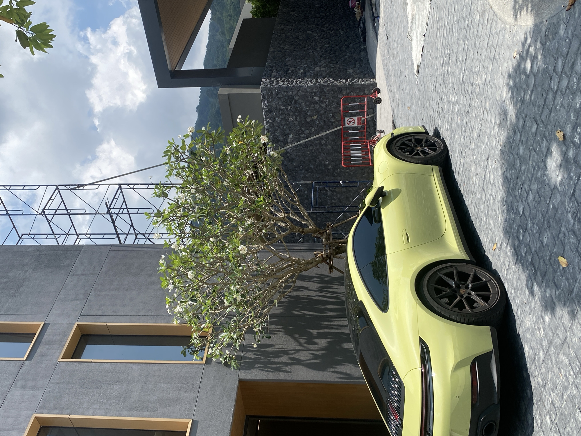 A light yellow (or lime green) Porsche sports car is parked on a textured grey paved surface, angled towards the right, under a clear blue sky with white clouds. The car, which appears to be a 911 series, is illuminated by strong sunlight, casting sharp shadows on the ground and adjacent building. Its rear wheel, brake caliper, and exhaust pipe are visible, along with a partial view of its sleek profile.
To the left of the car stands a modern building characterized by dark grey, textured walls and rectangular windows framed with light-colored borders. Above this building, a large, intricate metal scaffolding structure, possibly for a billboard or ongoing construction, is visible against the sky. A prominent tree, likely a Frangipani or Plumeria, with white blossoms and green leaves, stands between the car and the building, creating dappled shadows on the wall.
In the background, a distant mountain range can be seen. Near the parked car, red and white safety barriers are positioned, and one prominently features a circular red sign with a white graphic of a dog inside a red circle with a diagonal line, indicating "No Dogs Allowed" or "No Pets." A stone-clad wall and another section of a building with a brown wooden soffit are also visible.
The scene depicts a bright, sunny day, likely midday given the strong shadows, in what appears to be an upscale outdoor environment, potentially a resort or modern residential complex in Choeng Thale, Thailand.