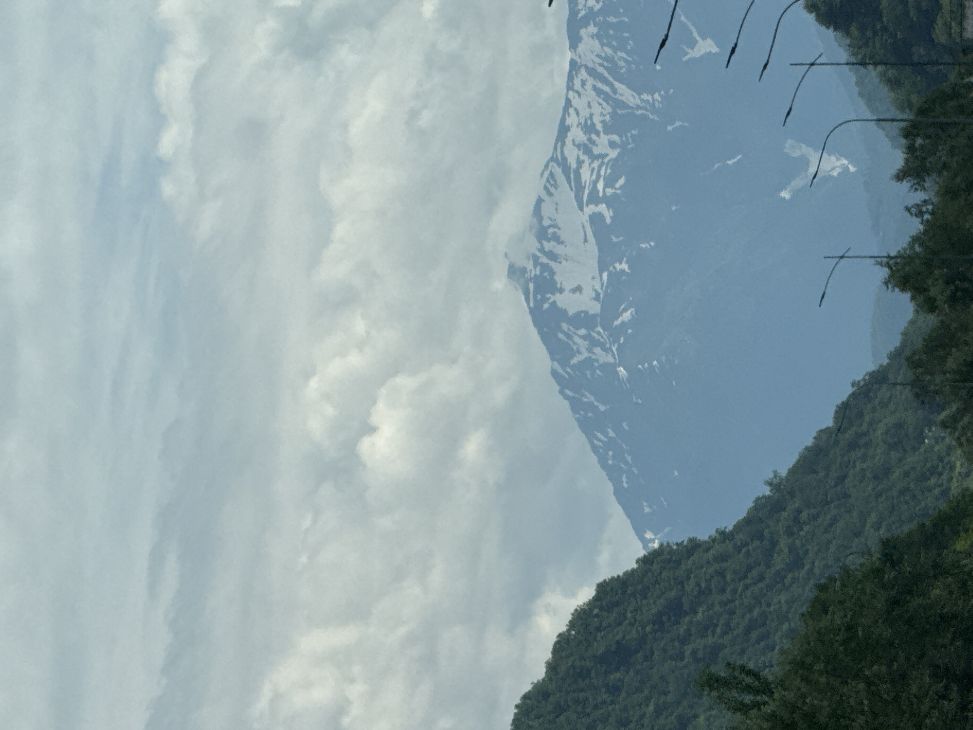 The image shows a panoramic view of a mountainous landscape with a partially snow-capped peak in the distance. The foreground features a steep, tree-covered mountainside on the left, with dark green foliage. In the middle ground, the imposing mountain range dominates the scene, its slopes streaked with snow. The sky above is filled with a mix of white and grey clouds, suggesting overcast conditions. Several streetlights with curved poles are visible in the lower right portion of the image, indicating the presence of a road or urban infrastructure near the natural landscape. The overall impression is one of natural beauty and a blend of rugged wilderness with hints of human development. The location is identified as Kepsha, Russia, a village in the Lazarevsky District of Sochi, known for its mountainous terrain and proximity to the Black Sea.