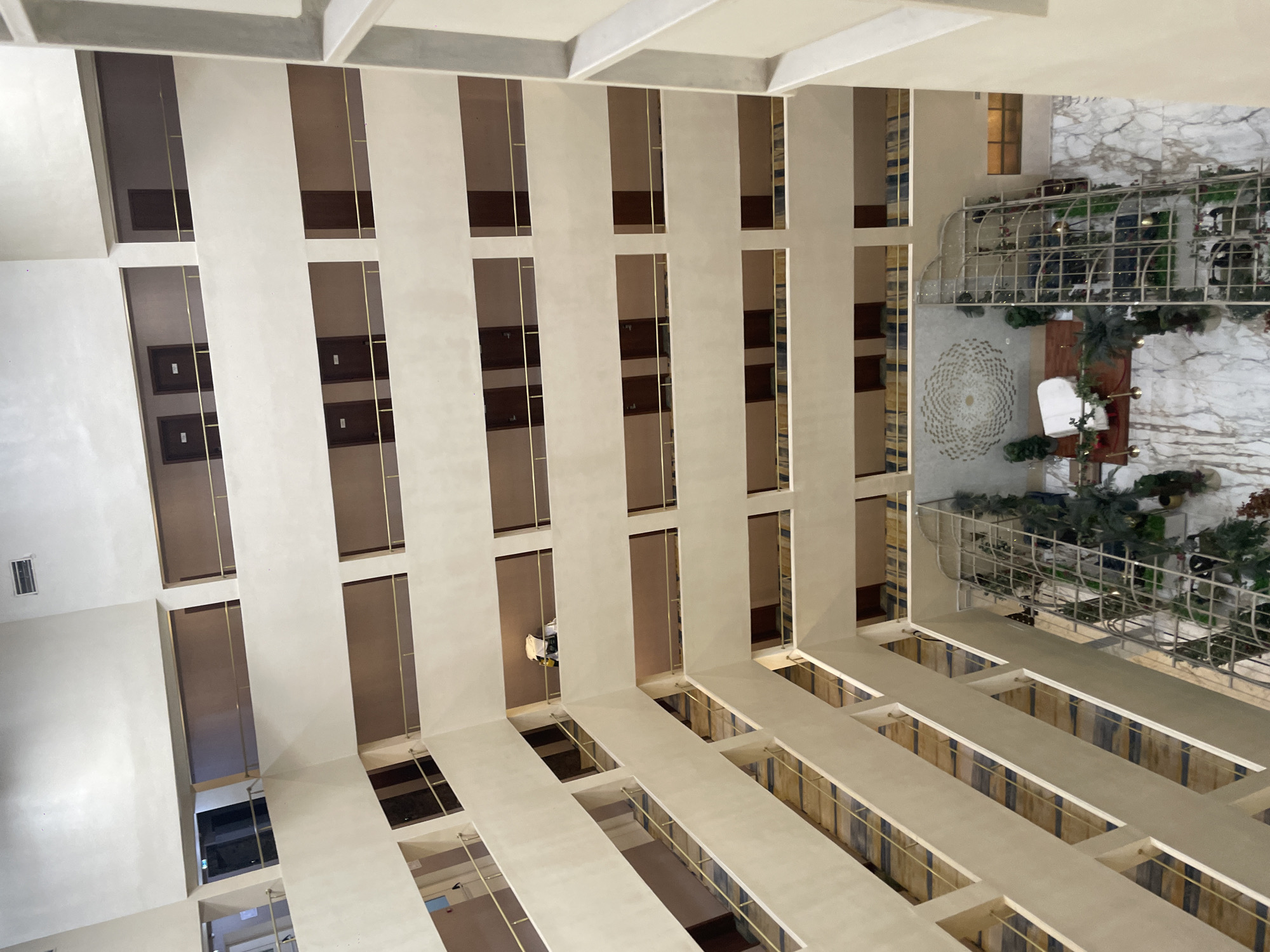 The image shows the interior of a multi-story atrium, likely a hotel or office building. The atrium features multiple levels of balconies with rooms or offices facing inwards. The architecture is modern and sleek, with beige walls and gold railings. On the ground floor, there is a decorative carpet, some plants, and what appears to be a white piano. The scene is well-lit, suggesting interior lighting. There are no people clearly visible in the image. The location of Oskemen, Kazakhstan is not indicated by any visible elements in the image.