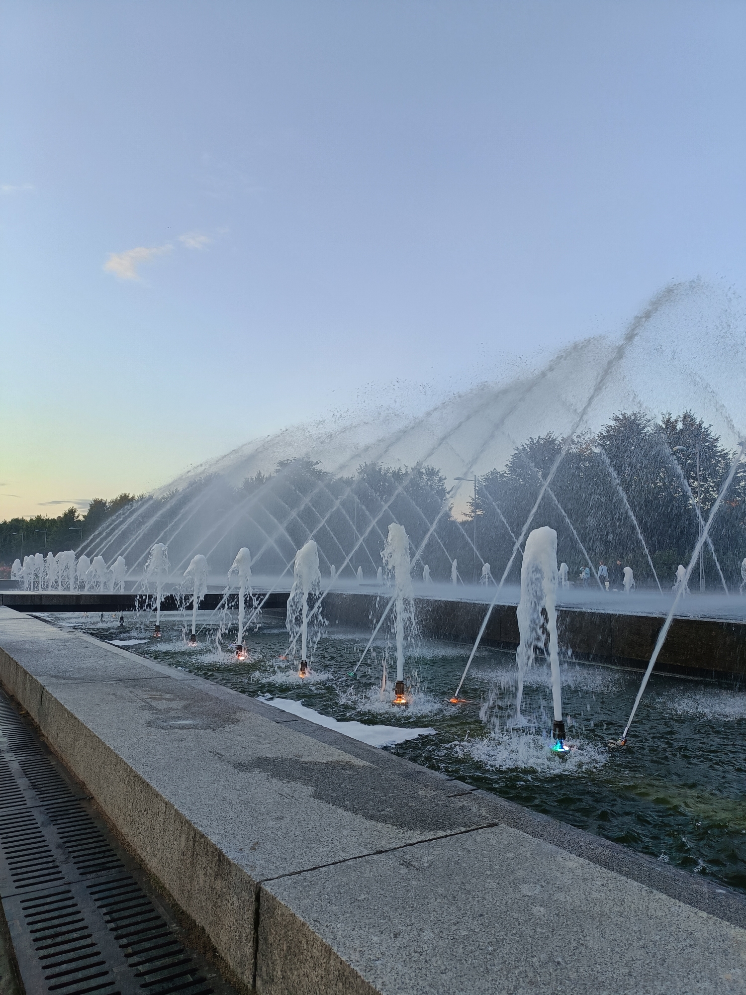 The image depicts a large, multi-tiered fountain system in the city of Saint Petersburg, Russia. The fountain is in operation, with numerous jets of water arcing upwards and across the scene, creating a dynamic and ethereal display. In the foreground, a stone pathway with a metal grate runs along the left side of the image. The fountain itself consists of rows of vertical water spouts, some of which are illuminated with colored lights, casting a subtle glow on the churning water below. In the background, a line of lush green trees forms a backdrop, with streetlights visible among them. The sky is a pale blue, suggesting it is either early morning or late evening. A few faint clouds are visible. No people are clearly discernible in the image, though there may be figures further back within the park setting. The overall impression is one of a public park or recreational area, enhanced by the striking water feature.