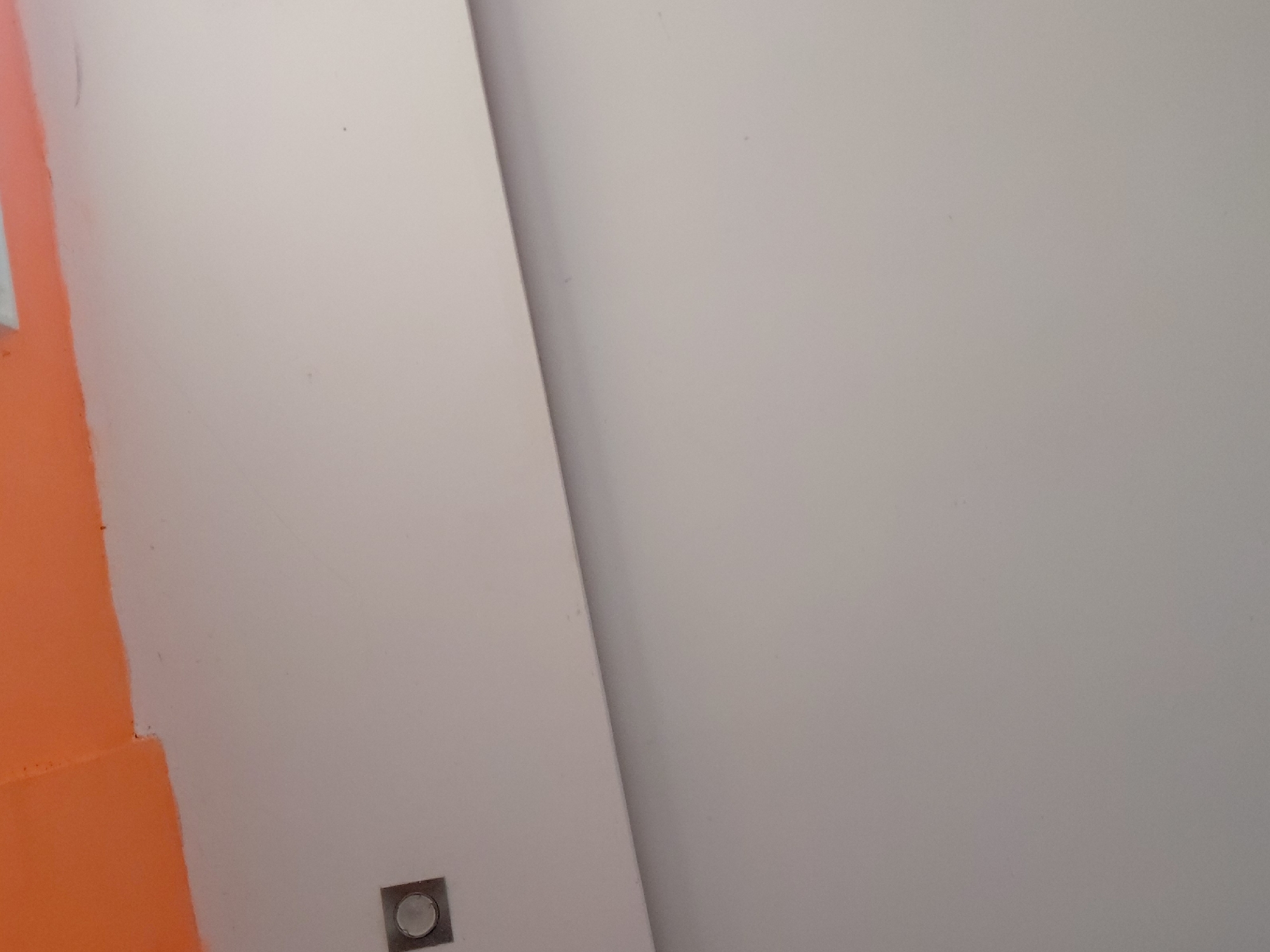The image displays an abstract indoor scene, primarily featuring two distinct wall sections. On the far left, a bold, solid orange surface extends vertically and diagonally, suggesting a corner or a painted partition. This orange section contrasts sharply with the main light gray or off-white wall surface that dominates the central and right portions of the frame. A prominent, darker diagonal line runs across the image from the upper-center towards the bottom-right, dividing the light-colored surface into two planes, possibly indicating a corner, a seam between two panels, or a significant shadow.
Near the bottom-center of the light gray wall, a small, square-shaped fixture is visible. It has a metallic or dark gray frame surrounding a recessed circular center, which could be an electrical outlet, a light switch, or a minimalist light source. The wall surfaces appear smooth with a matte finish. There are no people present, and no specific activity is taking place. The scene suggests an interior space, potentially in a modern building or an area undergoing renovation, given the clean lines and contrasting colors. No text is visible in the image. The setting is Santa Ana, Venezuela.