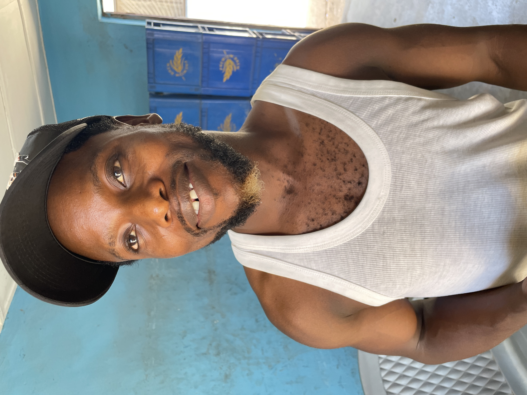 The media file displays a headshot and upper torso of a man in a casual, indoor setting in Jos, Nigeria. The man, who has a dark complexion, is looking directly at the camera with a slight, friendly smile, revealing his teeth. He has a short, dark beard with a distinct patch of blonde hair on his chin, and a mustache. He is wearing a black baseball cap backwards and a white, ribbed tank top. Visible on his chest are small, dark hair follicles or spots.
The background features a vibrant blue wall. To the right side of the frame, several blue plastic crates are stacked. Each crate bears a prominent yellow logo, which appears to be a stylized sheaf of wheat or a similar grain design, possibly accompanied by unreadable text like "PREMIUM". These crates suggest the setting might be a storage area, a shop, or a local establishment like a bar, likely for beverages. The lighting is bright and even, indicating that the photo was taken during the day. The overall atmosphere is relaxed and informal.
