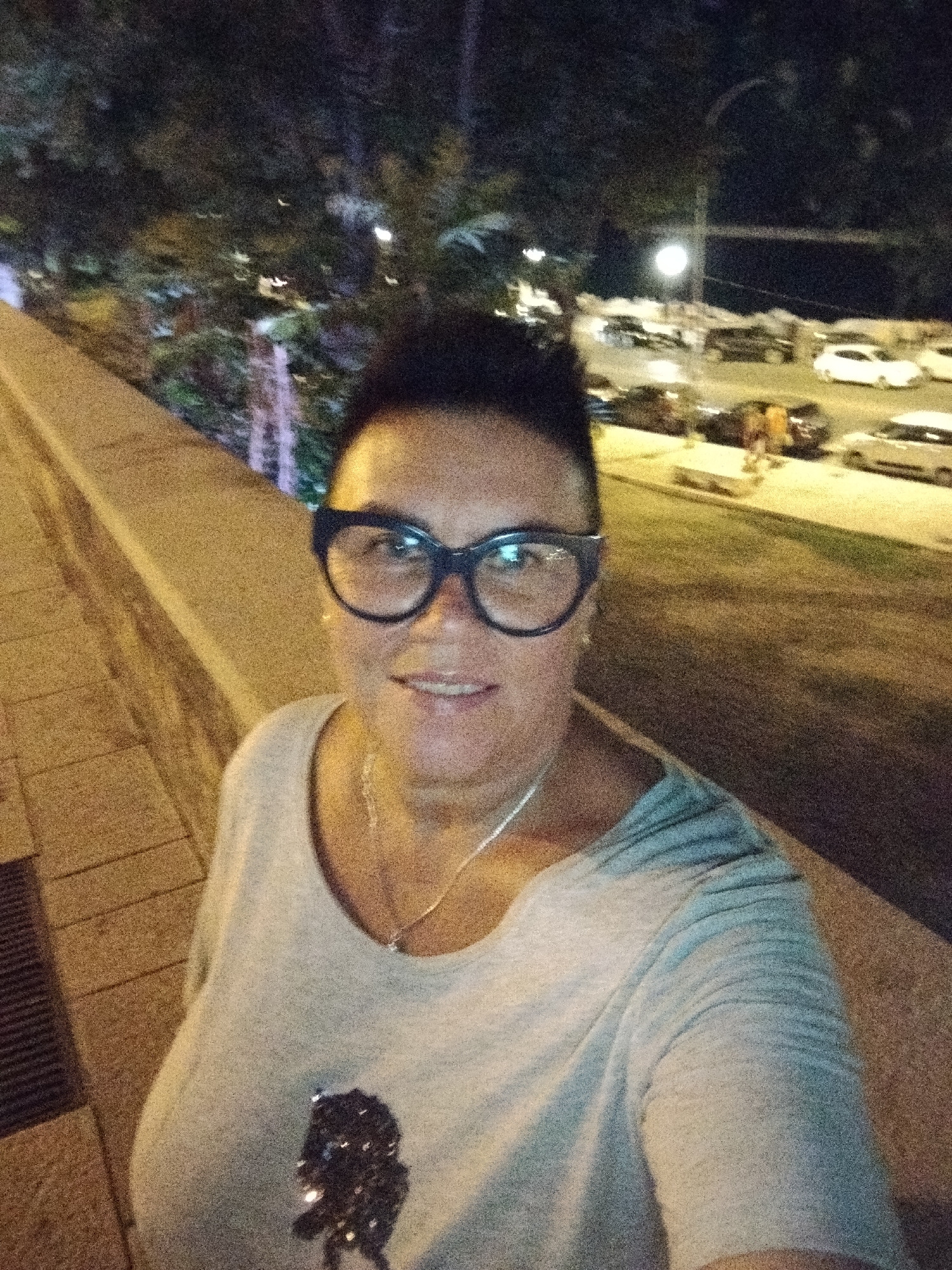 In a nighttime outdoor setting, a woman wearing large black-rimmed glasses and a light gray t-shirt with a sequined design on the front is captured in a close-up shot. She is smiling and looking towards the camera. Behind her, a blurred background depicts trees and parked cars illuminated by artificial lights, suggesting an urban environment, possibly in the city of Bari, Italy. A concrete barrier runs along the left side of the frame.