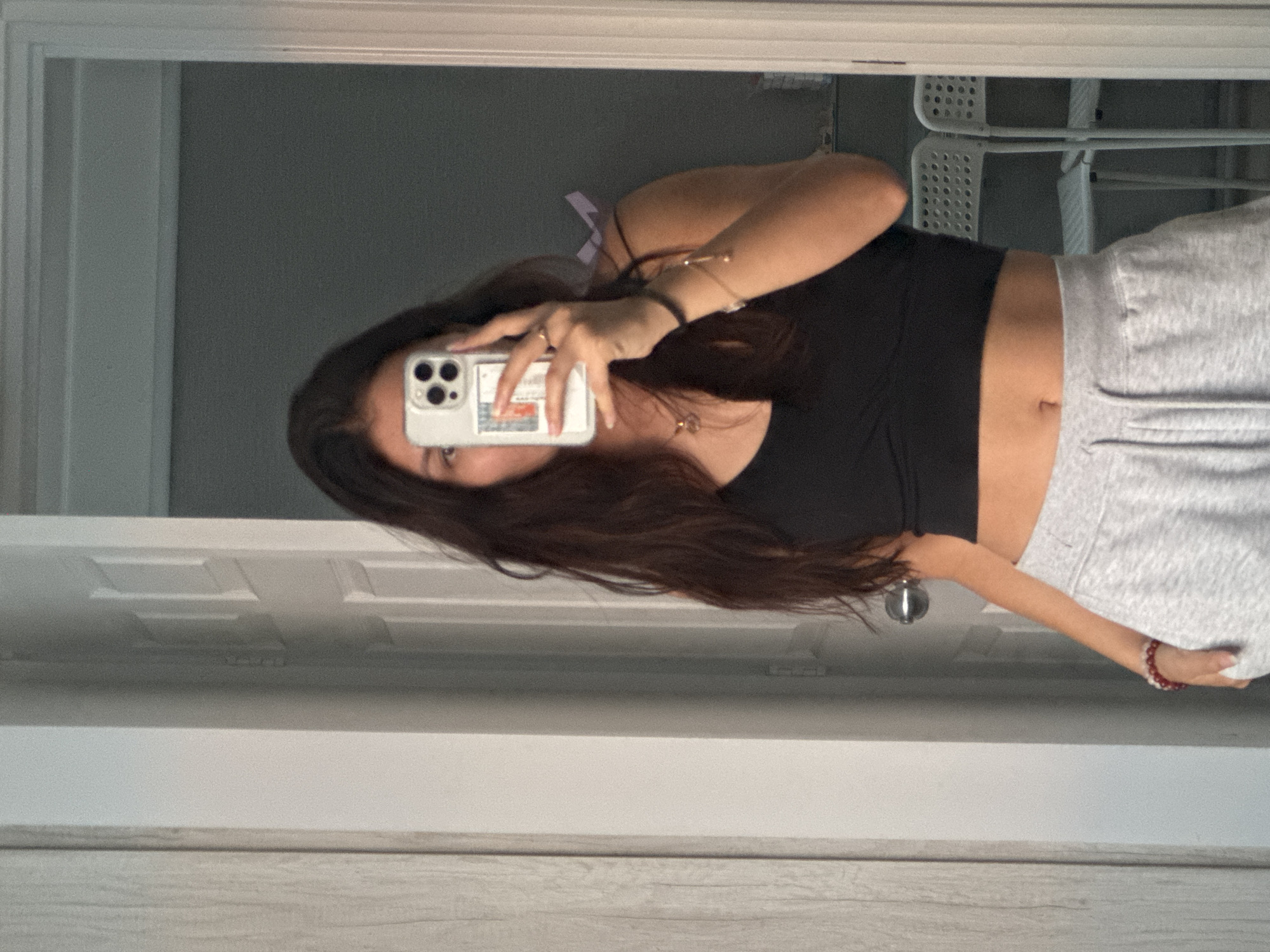 A woman is taking a selfie in a mirror. She is wearing a black top and gray sweatpants. Her long, dark hair is partially obscuring her face. She is holding a white smartphone with multiple cameras in her right hand. The reflection shows a white, metal chair in the background. The image appears to be taken indoors, possibly in a bedroom or bathroom. The location in the city of San Francisco Ocotlán (Ocotlán), Mexico, is not discernible from the image.