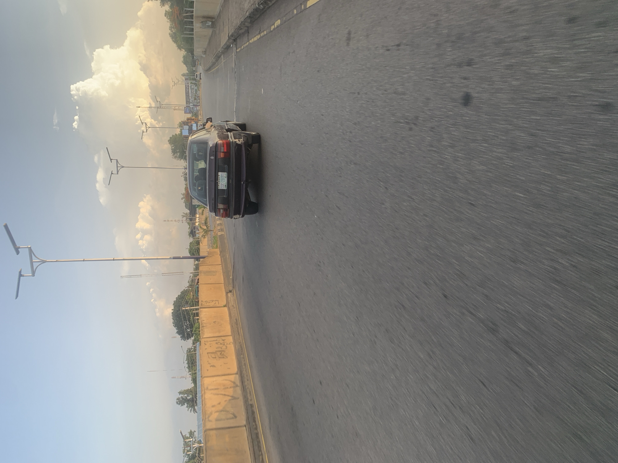 The image shows a view from a moving vehicle on an elevated road in Jos, Nigeria, at sunset. A dark sedan is in front, traveling in the same direction. The road has two lanes in this direction and a concrete barrier on the left. On the other side of the barrier, there are trees, buildings, and streetlights. The sky is a mix of blue and golden clouds, indicating it is late afternoon or early evening. The road surface is asphalt and shows signs of wear. There is a yellow line marking the edge of the lane on the right side of the road.