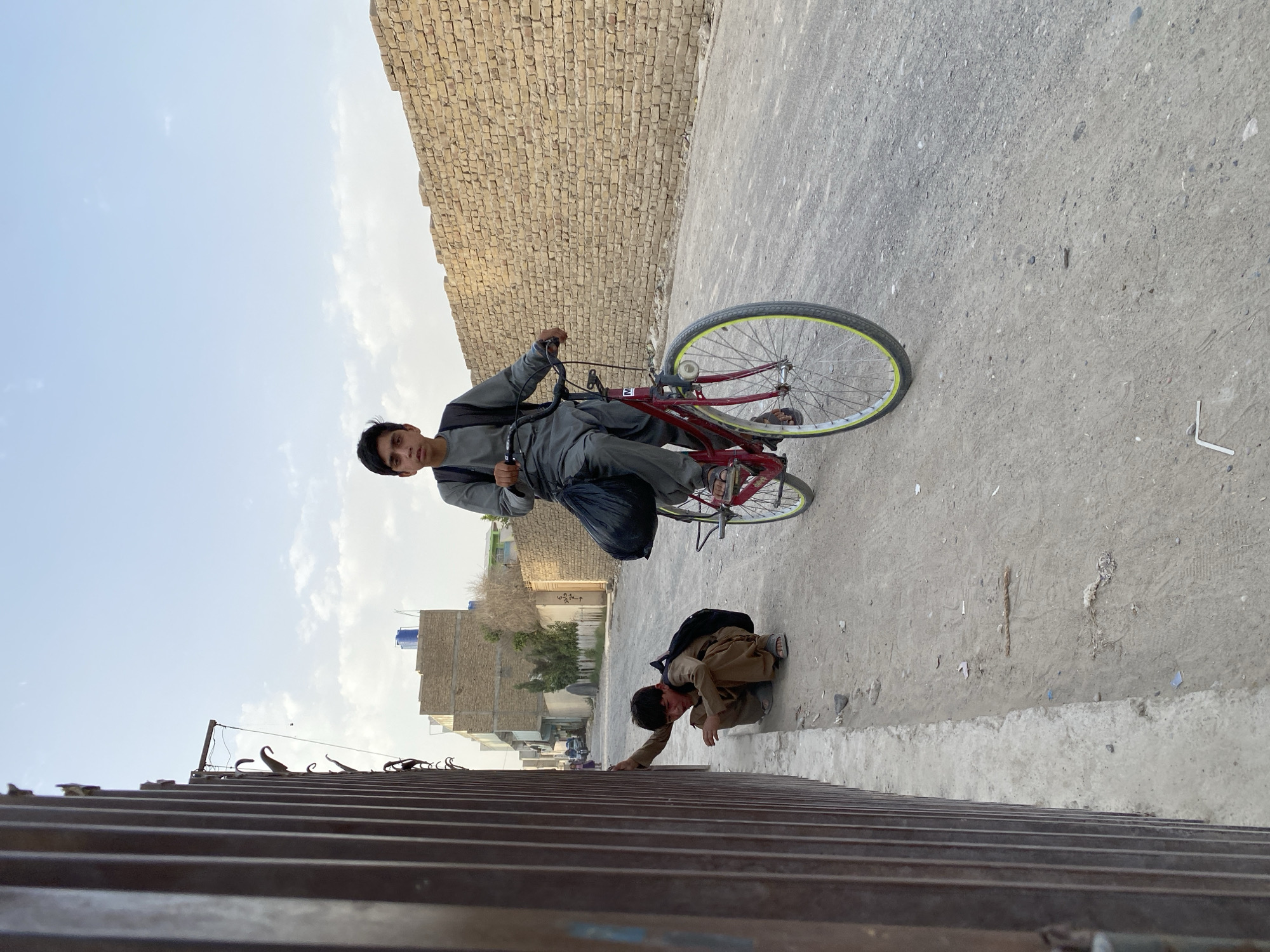 An outdoor, eye-level, medium shot shows two young boys in a dusty, urban environment under a clear, pale blue sky with scattered clouds. The boy on the left is crouching near a tall, dark metal fence, looking towards the ground with his right hand on the fence. He has dark hair and is wearing a tan outfit and a backpack. The boy on the right is seated on a red bicycle with a black bag slung over his shoulder, riding down a dirt road. He has dark hair and is wearing a gray outfit. In the background, there are buildings constructed of rough brick and some trees. The ground is made of dirt and gravel, with some scattered debris. The overall impression is one of everyday life in a dry, possibly arid, city. The scene appears to be in Kandahar, Afghanistan, given the architectural style and the typical clothing worn by the boys.