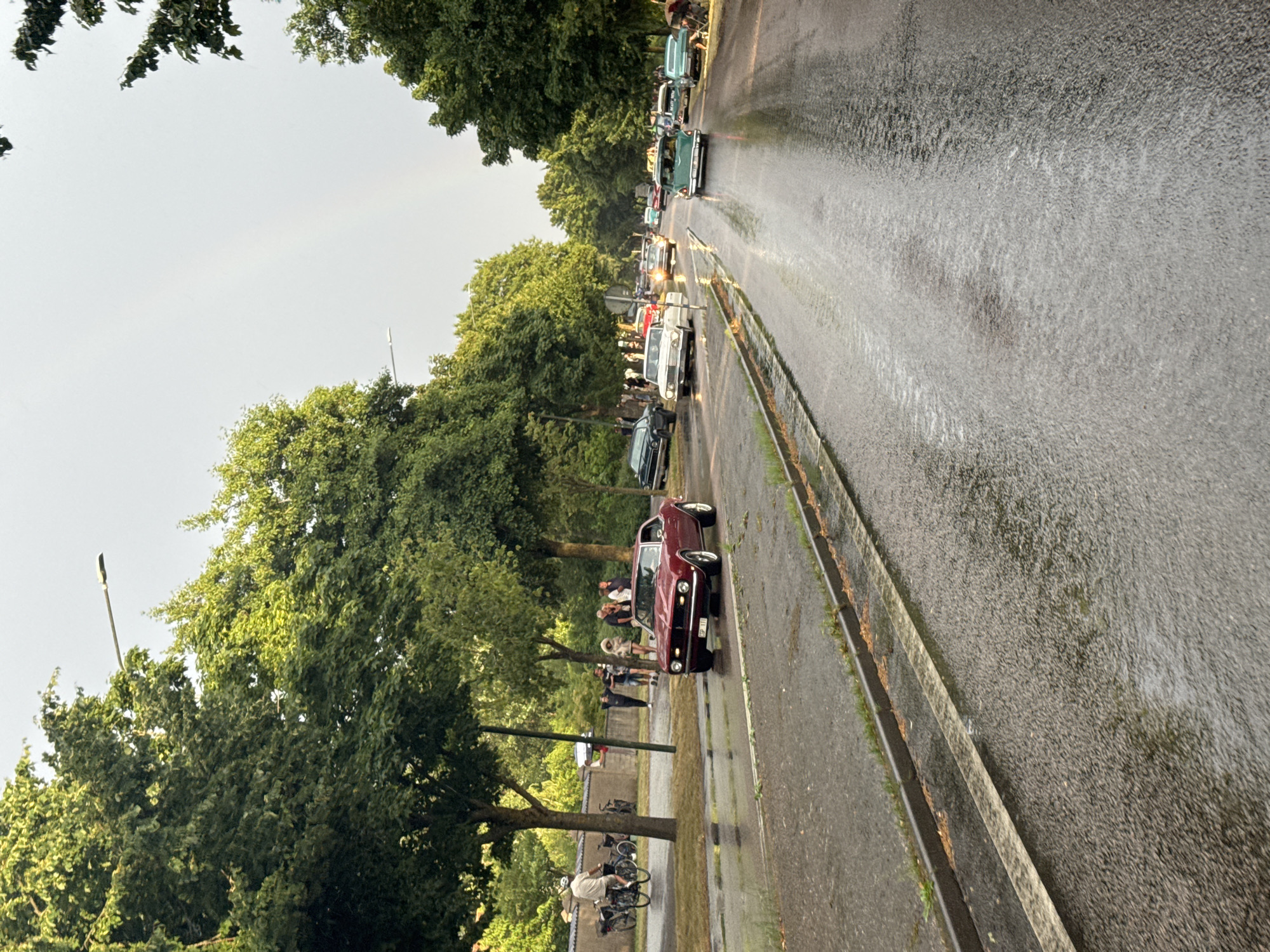 The image captures a street scene in Falkenberg, Sweden, under overcast skies with a faint rainbow visible. The road is wet, indicating recent or ongoing rain, with visible puddles and reflections. A procession of vintage cars is driving down the street. In the foreground, a dark red classic car, possibly a Chevrolet Camaro, is the most prominent vehicle. Further down the road, other classic cars, including a white one and a teal-colored one, are visible. Several people are gathered on the sidewalks and near the cars, some watching the vehicles, while others appear to be enjoying the event. Trees line the street, their foliage lush and green. A street light is also visible. On the left side of the road, a person on a bicycle is seen near parked bicycles. The overall atmosphere suggests a car show or a classic car rally taking place in a residential or tree-lined urban area.
