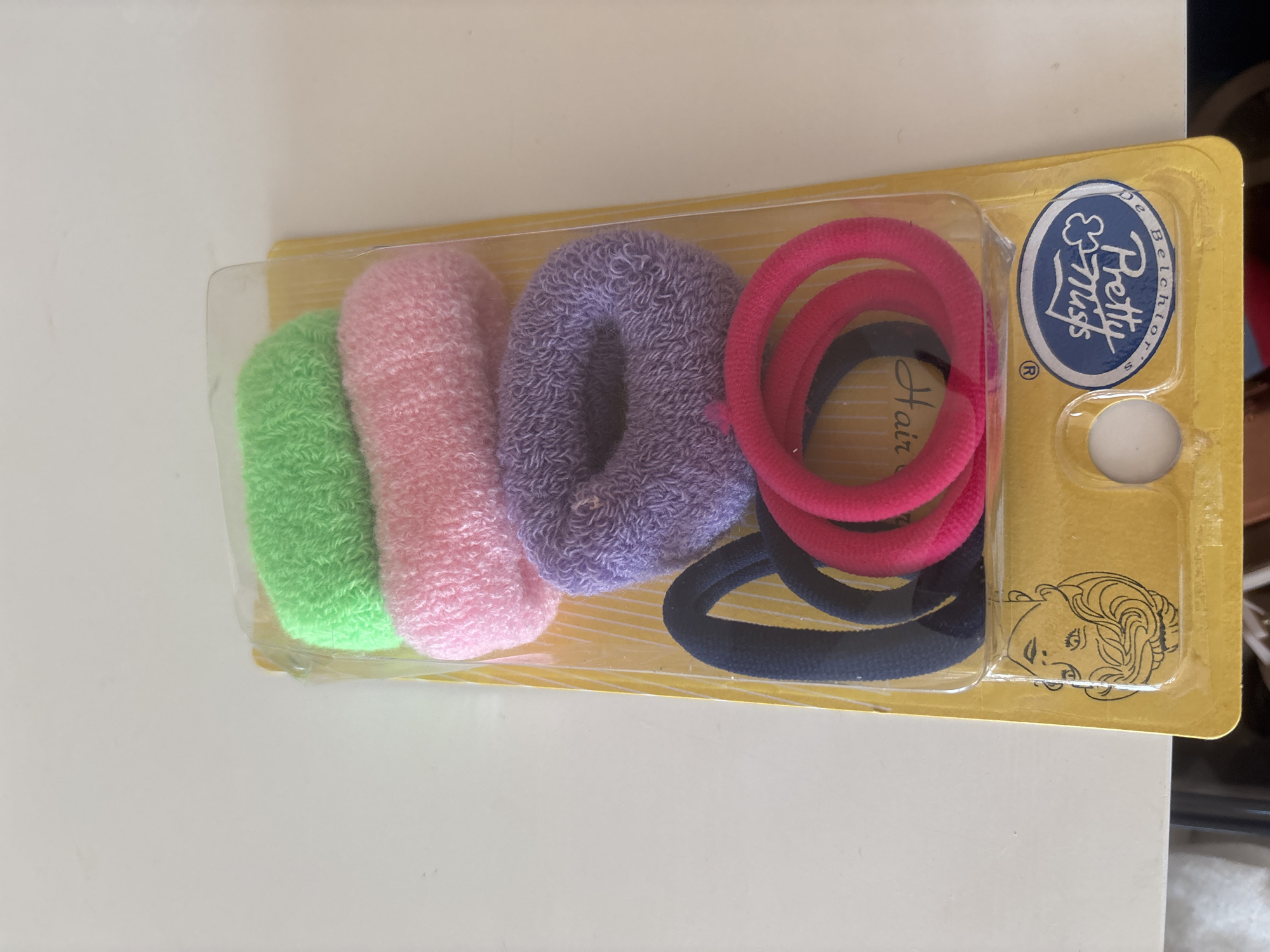 The image shows a retail package of six hair ties on a white surface, likely a table, indoors. The packaging is clear plastic on a yellow cardboard backing. Inside, there are three fabric-covered hair ties stacked vertically: a green one at the top, a pink one in the middle, and a purple one at the bottom. Below these, there are three more hair ties arranged horizontally: a dark blue one, a bright pink one, and another bright pink one slightly overlapping the first. The yellow backing has a graphic of a person's head with curly hair and a beard, along with text and a circular logo that reads "Pretty Ties by Belchior's". The background is a plain white wall. There is no indication that this image was taken in Dubai, United Arab Emirates.