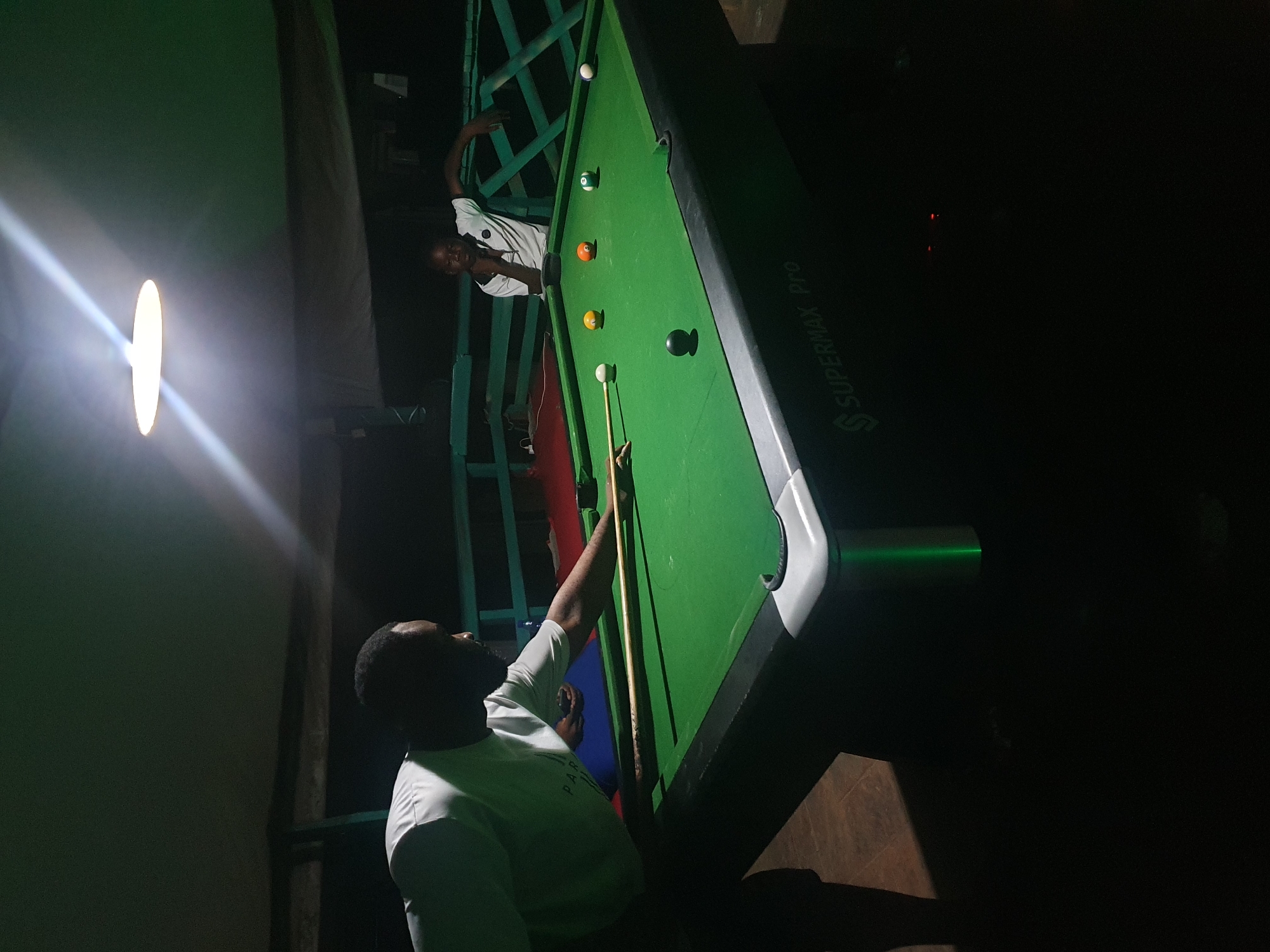 Two individuals are playing billiards in an outdoor, covered area at night in Lokoja, Nigeria. The scene is dimly lit by an overhead light fixture. A man in a white t-shirt is actively playing, leaning over the green billiard table, cue in hand, aiming at a white ball. Several other billiard balls are scattered across the table, including a black ball, a yellow ball, an orange ball, and a striped ball. To the right, another person, a young man in a white t-shirt with black accents, leans against a green railing, observing the game. The table itself is a "SUPERMAX Pro" model. The environment appears to be a recreational space, possibly a bar or a private establishment, with wooden structures and a railing system. The ground is a dark, textured surface, possibly wood or concrete.