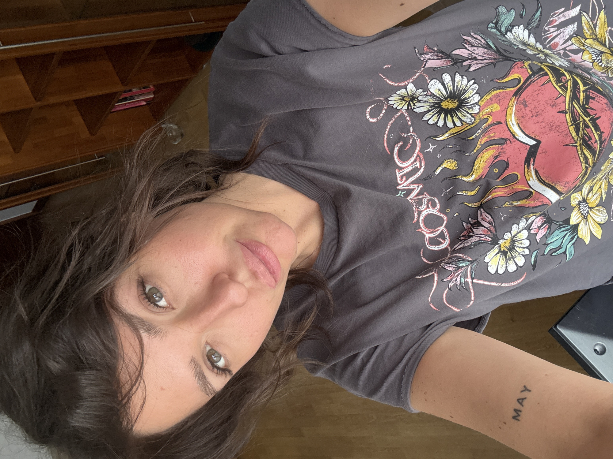 A woman is lying on a wooden floor, wearing a gray t-shirt with a heart, flames, thorns, and flower design. "MAY" is tattooed on her right forearm. A wooden bookcase with objects is visible behind her. The location cannot be determined.