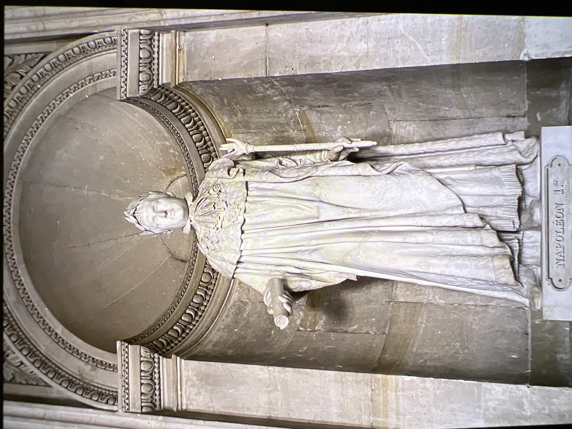 The image shows a full-body marble statue of Napoleon Bonaparte. He is wearing a laurel wreath on his head, a ceremonial robe draped over his shoulders, and a long tunic underneath. His right hand holds a scepter or staff, and his left hand holds a scroll. The statue is positioned in a semicircular niche within a stone architectural setting. The niche has decorative molding around its arch and pilasters on either side. A marble pedestal supports the statue, with the inscription "NAPOLEON 1ER" clearly visible on its front. The statue is white and appears to be well-preserved, with intricate details in the drapery of his clothing and the facial features. The background is composed of rough-hewn stone blocks.
This statue is located in the city of Verona, Italy. Specifically, it is found inside the Loggia del Consiglio, a Renaissance loggia that is part of the Palazzo Barbieri, which serves as the city hall of Verona.