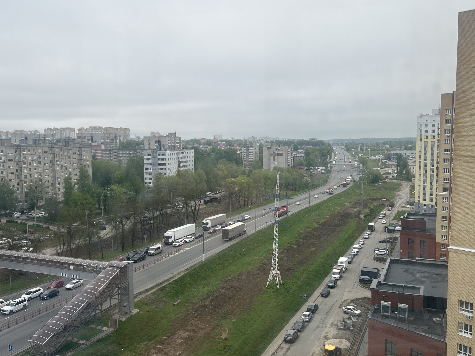 The image depicts a cityscape on an overcast day, possibly in Unknown, Unknown. The foreground includes a pedestrian bridge over a multi-lane road with moderate traffic consisting of cars and trucks. Beside the road are grassy areas with an electric power transmission tower. Adjacent to the grassy area is a smaller road lined with parked vehicles. Beyond the road are low-rise buildings and some taller apartment complexes. The sky is uniformly gray, indicating overcast conditions.