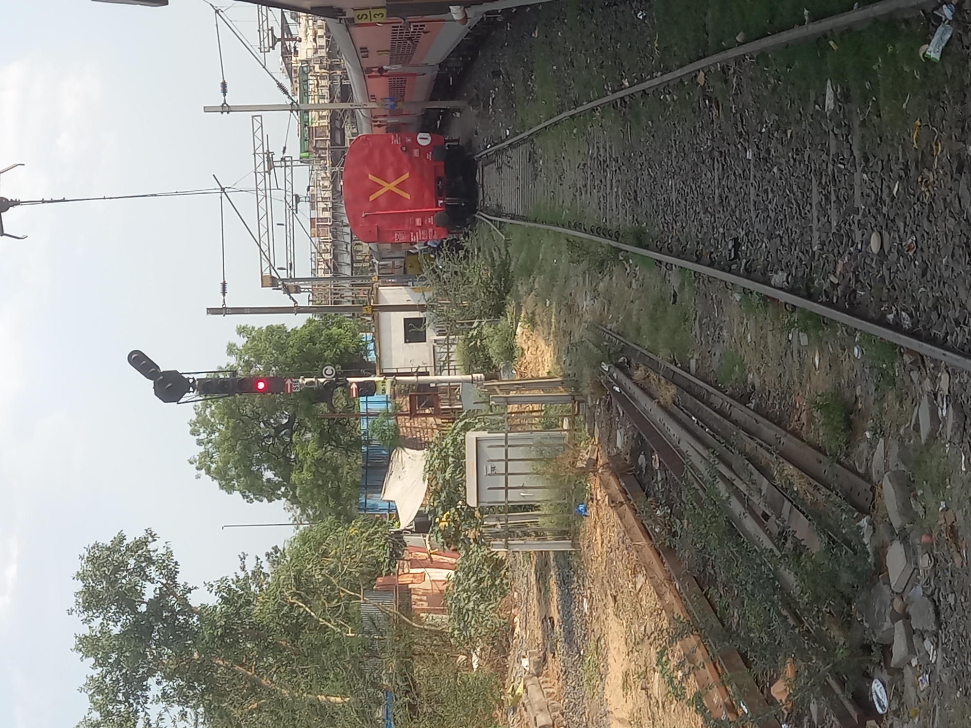 The image shows a railway scene in Bhopal, India. A red train car with a large yellow "X" is coupled to another car on a track that curves away from the viewer. Overhead electrical lines and signal equipment are visible. A signal light is showing red. Adjacent to the tracks are ballast, vegetation, and discarded materials. A small white building and partial views of other structures are visible behind the tracks. Discarded rails are stacked in the foreground.