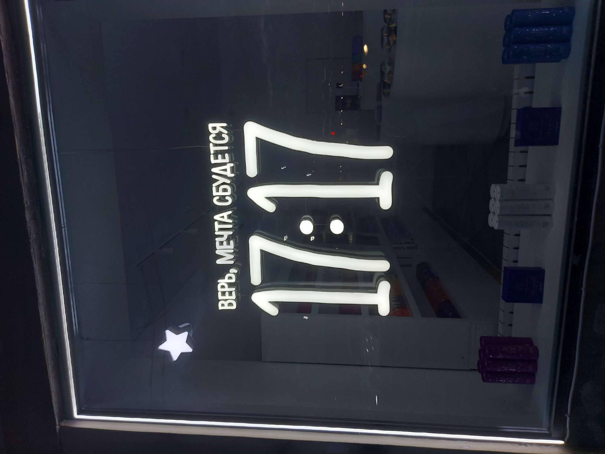 The image shows a store display behind a glass window. In the center is a neon sign displaying "11:11" vertically, with the Russian text "ВЕРЬ, МЕЧТА СБУДЕТСЯ" (Believe, a dream will come true) alongside. To the right of the sign, there are several items stacked on shelves, appearing to be rolls or packaged items in various colors including blue, gray, and purple. A white star is below the text and left of the sign.
The items on the shelves and the overall aesthetic suggest a retail environment, possibly a store selling stationery, arts and crafts, or similar goods. The image likely originates from Saint Petersburg, Russia due to the Russian text on the neon sign.