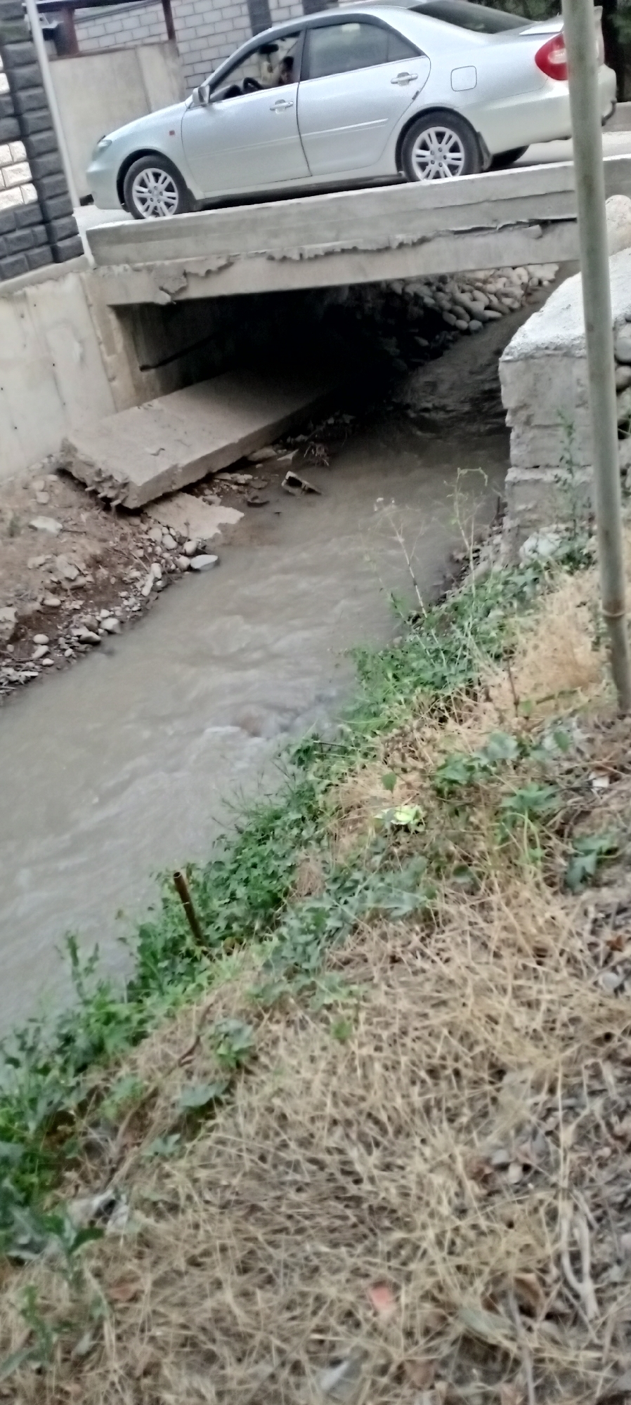A silver sedan is parked on a concrete bridge over a dirty water channel. The channel is lined with concrete on the left bank and natural earth with vegetation on the right bank. The bridge appears to be made of concrete with visible signs of wear and tear, with a broken section on the left side. The water in the channel is murky brown and appears to be flowing. The surrounding environment consists of a brick wall and a paved area on the left, and dry grass and sparse vegetation on the right. A partial view of a person is visible in the driver's seat of the car. The scene is outdoors, under what appears to be daylight conditions. The location is identified as Sokuluk, Kyrgyzstan.