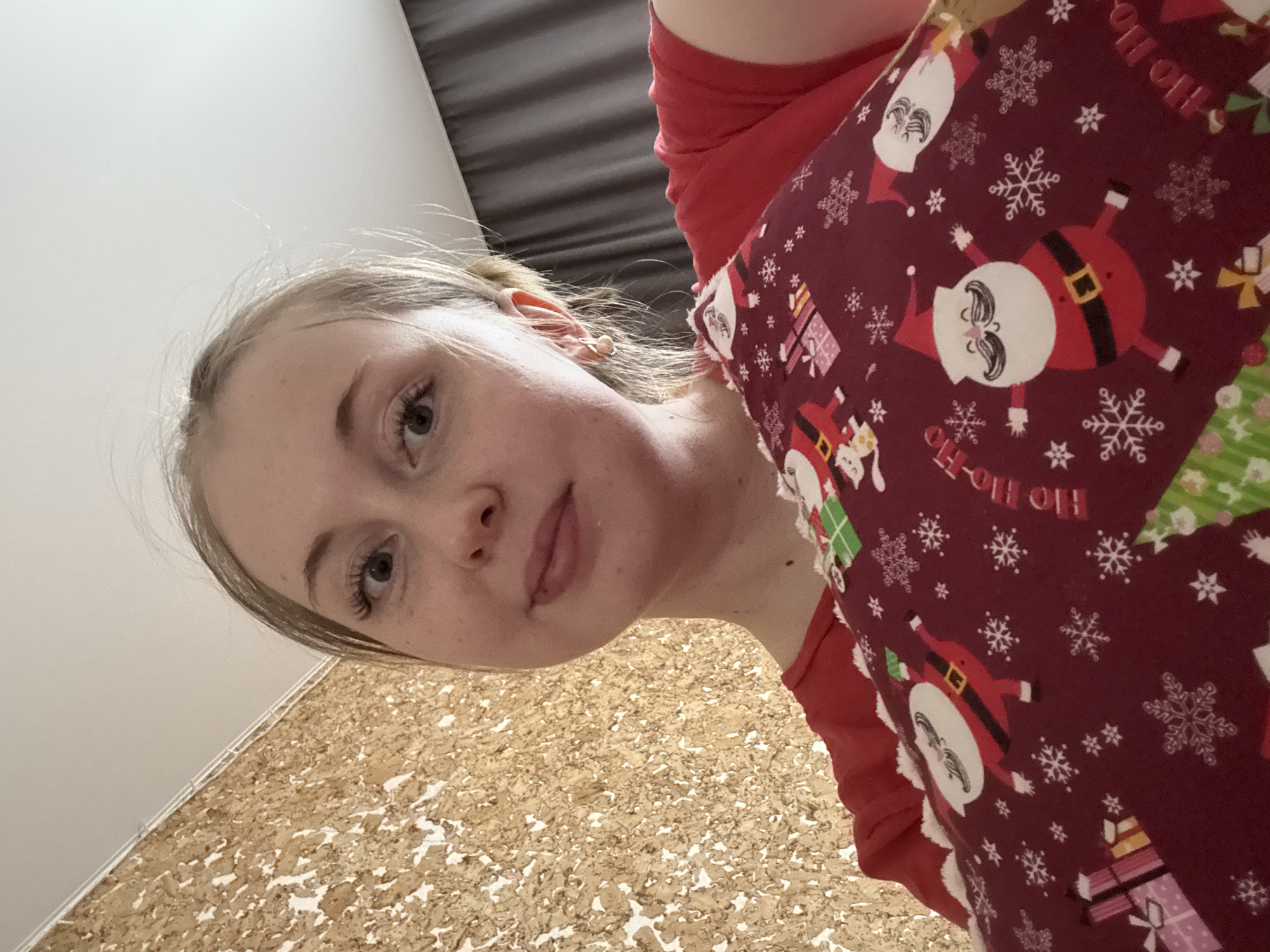 This image is a close-up, angled portrait of a young person in an indoor setting.
**Subjects:** The primary subject is a young individual with light blonde or light brown hair, which appears to be pulled back. Their face is visible, showing light-colored eyes looking directly towards the camera, a small nose, and closed lips with a neutral expression. They have fair skin, with a few visible freckles. The person is wearing a red long-sleeved top.
**Objects:** Over the red top, the person is wearing or holding a piece of fabric with a distinct Christmas-themed pattern. This pattern is predominantly dark red/maroon and features cartoonish Santa Claus figures dressed in red suits with white beards and hats. Various white snowflakes and wrapped gift boxes in different colors (pink, light blue, green) are scattered across the fabric.
**Visible Text:** The text "HO-HO" is visible in white lettering on the patterned fabric, integrated into the design.
**Scene/Setting:** The background indicates an indoor location. To the upper left, a plain white wall or ceiling is visible. To the upper right, there's a dark curtain or blind with horizontal pleats or slats. The lower part of the image reveals a textured floor or wall covering, appearing to be made of light brown cork or a similar composite material, with patches of lighter white/beige. A white trim or molding runs along the edge where this textured surface meets the white wall.
**Activity:** The person appears to be posing for a photograph, likely a selfie, given the close-up and angled perspective from above.
**Notable Details:** The Christmas-themed pattern on the fabric is a prominent detail, suggesting the photo might have been taken during the holiday season or features festive attire. The lighting is soft and even, typical of indoor environments. The overall mood is calm and casual.