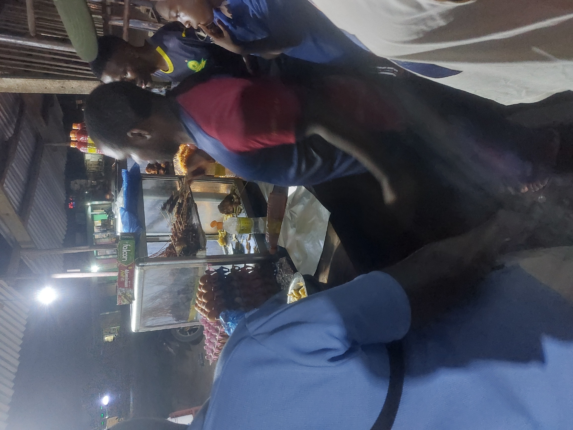 The image shows a food stall in Dar es-Salaam, Tanzania. Three men are visible around the stall. One man is in a red and blue shirt, another wears a blue shirt, and a third wears a blue and yellow shirt. The stall contains various food items, including sausages and skewered meats. Packaging for food products like "Falcon Aluminum Foil" is visible on a shelf. The location appears to be outdoors and at night, with streetlights illuminating the scene.
