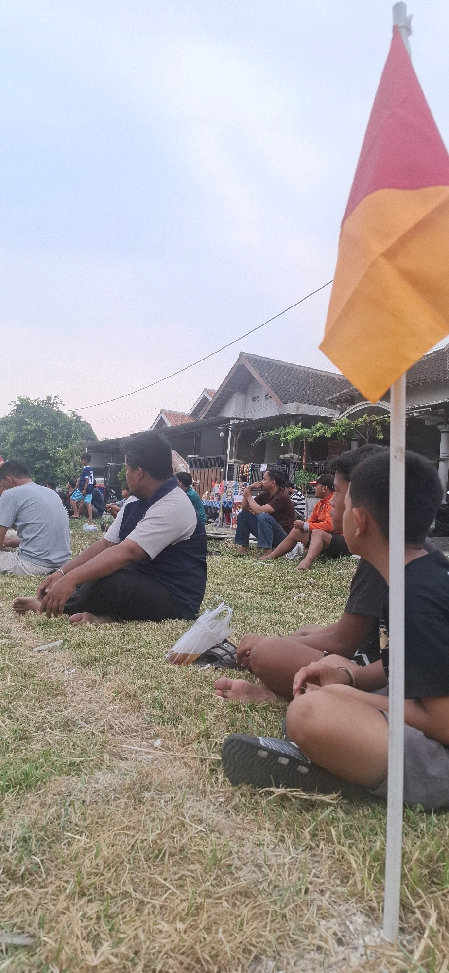In Desa Dibal, Indonesia, a group of people are sitting on dry grass, facing away from the camera. In the foreground, two young males are seated close together, with one wearing a black t-shirt and the other a grey shirt. Behind them, a man in a navy and grey shirt is also seated, with a plastic bag containing liquid to his right. Further back, other individuals are scattered across the grass, some in casual attire. The background reveals residential buildings with tiled roofs, and lush green trees. A flag, partially visible and made of red and orange fabric, stands on a white pole to the right of the frame, its color suggesting it might be related to a local event or organization. The sky above is a pale, overcast blue.