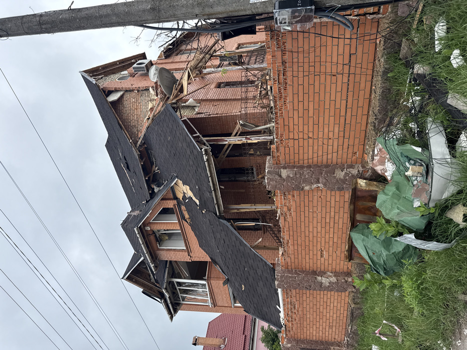 A severely damaged brick building, possibly residential, exhibits significant destruction. The roof is torn apart, windows are shattered, and parts of the structure appear collapsed or compromised. An electrical meter is visible on the exterior. Debris litters the area, including possible car wreckage covered with green tarps. The scene suggests a major incident, such as an explosion or impact, leaving the building uninhabitable.