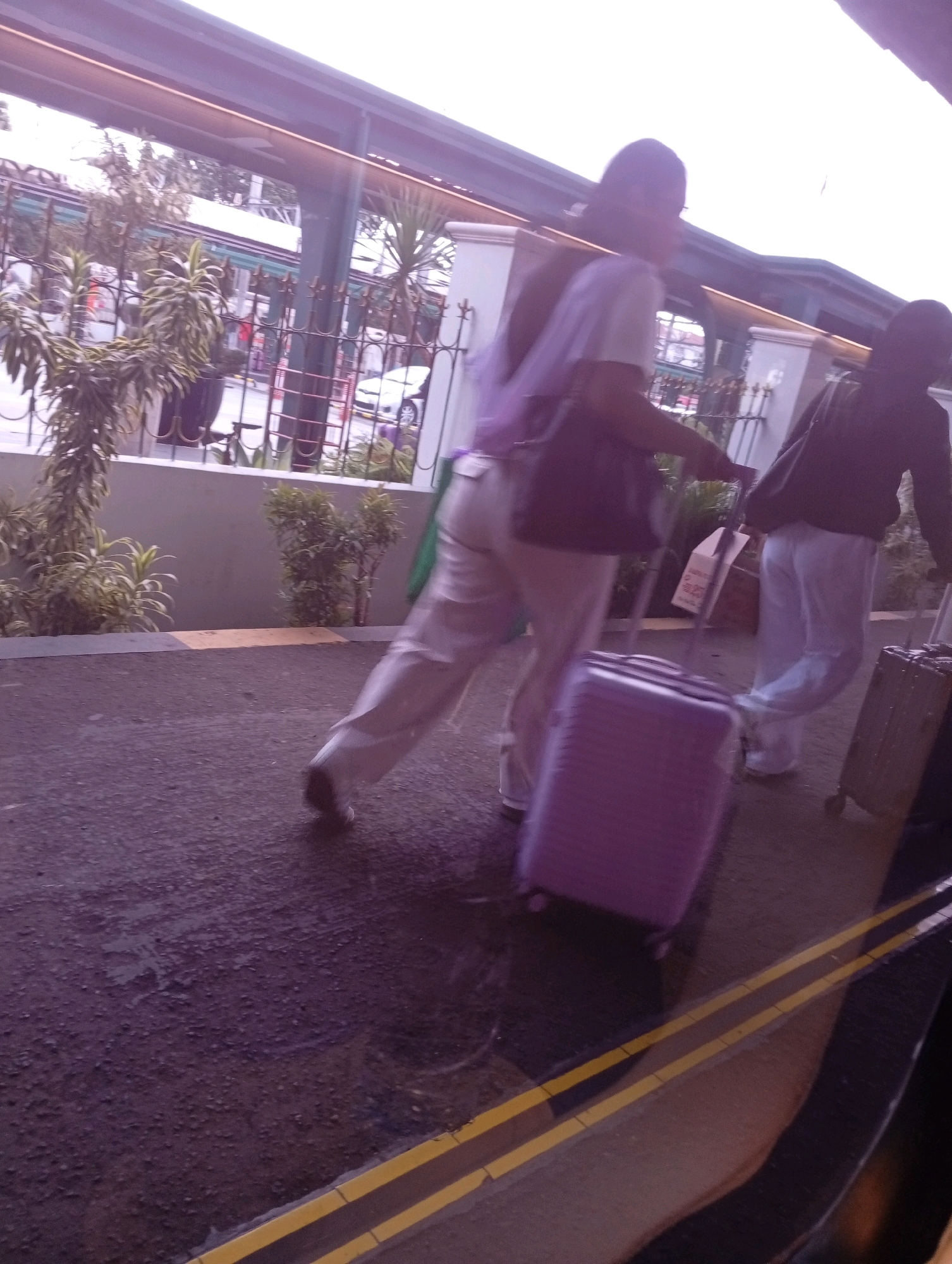 The image captures two individuals walking with luggage on a paved outdoor area, possibly a walkway or a station platform. The lighting suggests it is daytime, with some overexposure and glare.
The person in the foreground is wearing a purple backpack and light-colored pants, and is pulling a large purple suitcase with wheels. Their back is partially turned to the camera, and their face is obscured.
Behind them, another person is walking, wearing a dark top and light-colored pants, and pulling a darker suitcase. This person's face is also not clearly visible.
The environment includes some greenery in the background, a fence, and a building structure overhead with lights. The ground is a dark asphalt-like surface with yellow lines painted on it.
There is no specific visual information within the image that indicates the location as Yogyakarta, Indonesia.