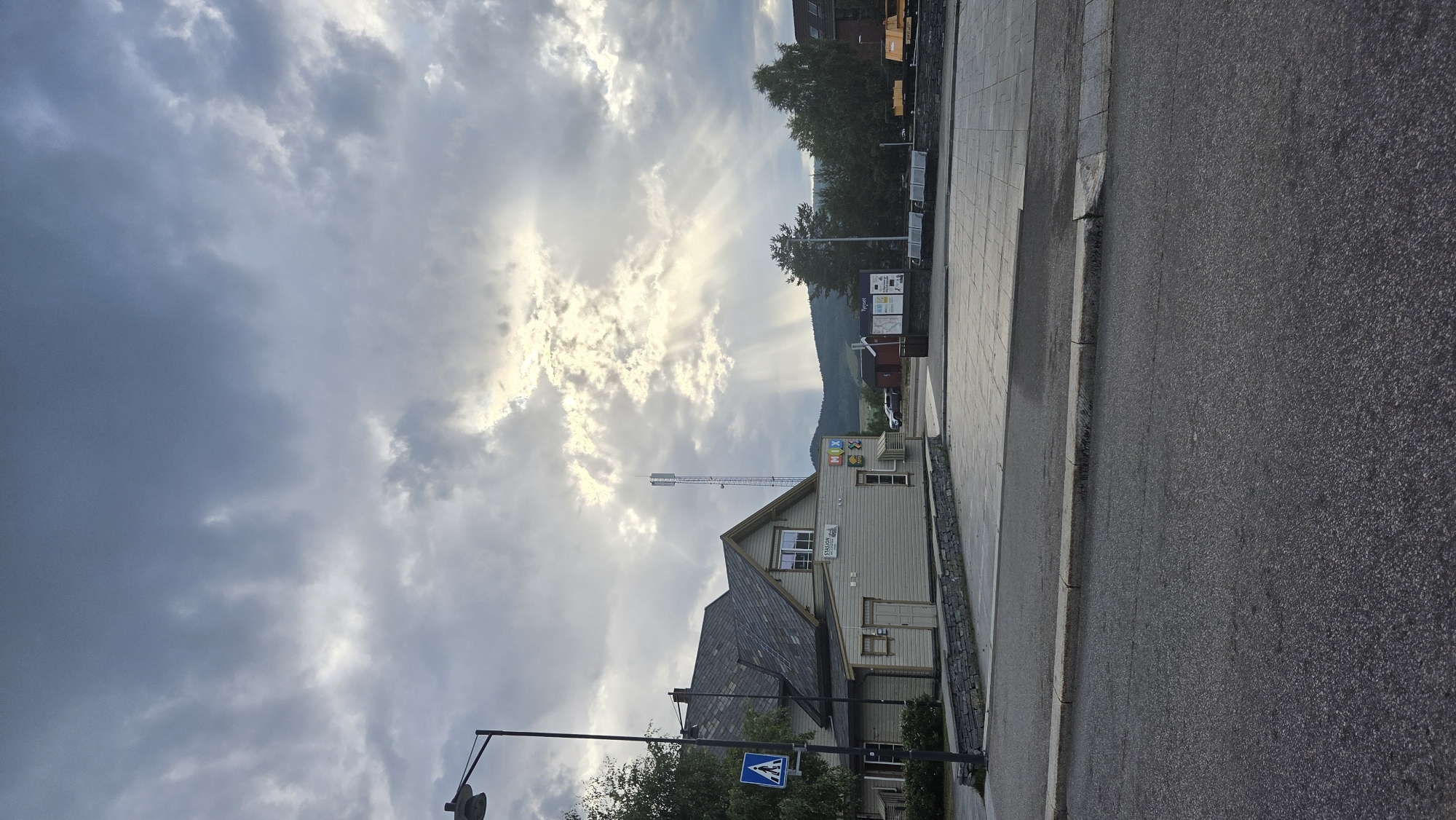 This is a photograph of the Tynset train station in Tynset, Norway, on a cloudy day. The sky is dramatic with sunbeams breaking through the clouds. The train station building is a light-colored wooden structure with a dark-tiled roof. A sign above a window on the building reads "STASJON". Next to the station is a blue and white pedestrian crossing sign. In the background, there is a telecommunications tower. The scene also includes a paved area in front of the station with benches and a map display. The foreground is a paved road. Trees and hills are visible in the distance. There are no people visible in the image.