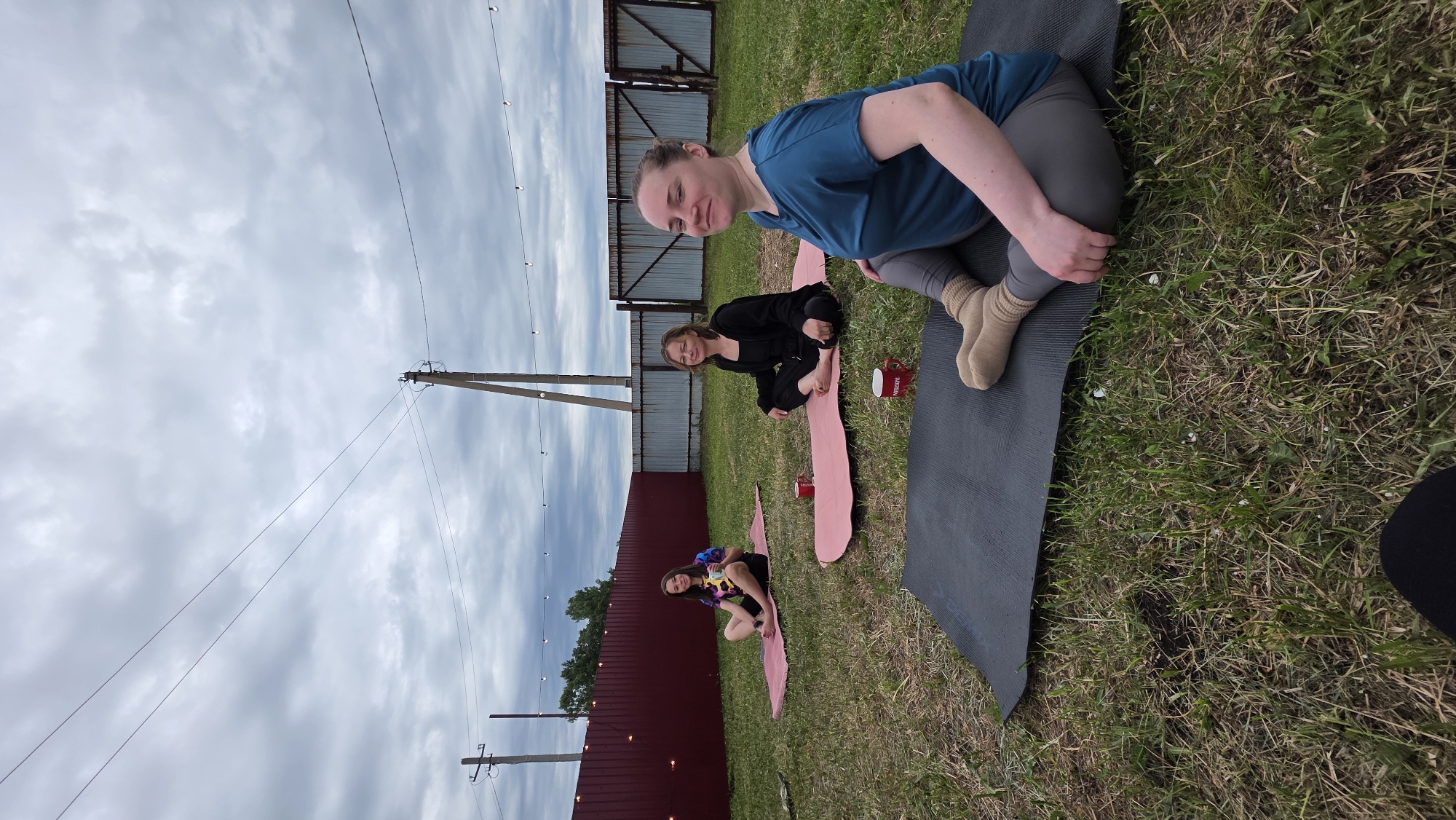 In Купоярово, Russia, three women are practicing yoga on a grassy backyard. Two women are seated on pink yoga mats, while the third is on a black yoga mat in the foreground. The woman in the foreground is wearing a teal t-shirt and grey leggings, and is in a seated yoga pose. The other two women are also seated in meditative poses. Two red mugs are visible on the yoga mats. A maroon fence runs along the background, with a metal gate to the right. Power lines and utility poles are visible in the cloudy sky.