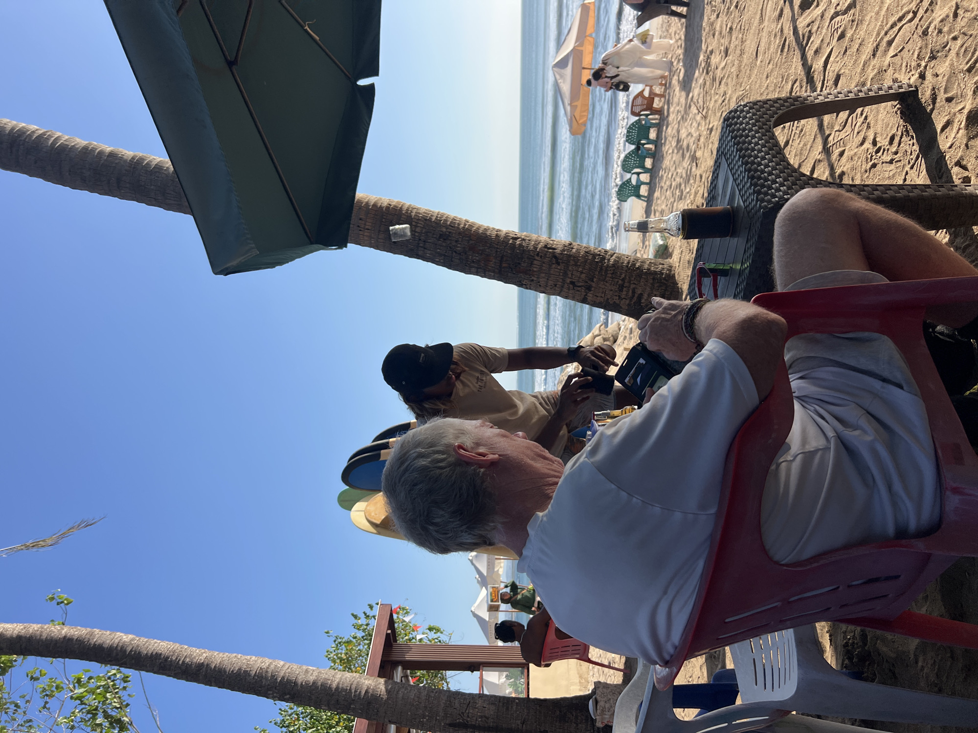 A man with graying hair sits in a red plastic chair facing away from the camera, looking at a tablet device on a black wicker table. Beside the tablet is a bottle of dark liquid. He is on a sandy beach in Kuta, Indonesia. Behind him, a man wearing a black baseball cap and a tan t-shirt with text on it is standing and looking down at something in his hands, possibly interacting with the man in the red chair. Two palm trees flank the scene, one on the left and one on the right, with a large green umbrella partially obscuring the right palm tree. In the background, the ocean with moderate waves can be seen, along with several colorful plastic chairs and two umbrellas on the beach. Further in the background, there are people walking and standing near a building that appears to be a beachside establishment. Several surfboards are propped up against a wooden structure to the left of the standing man. The sky is clear and blue.