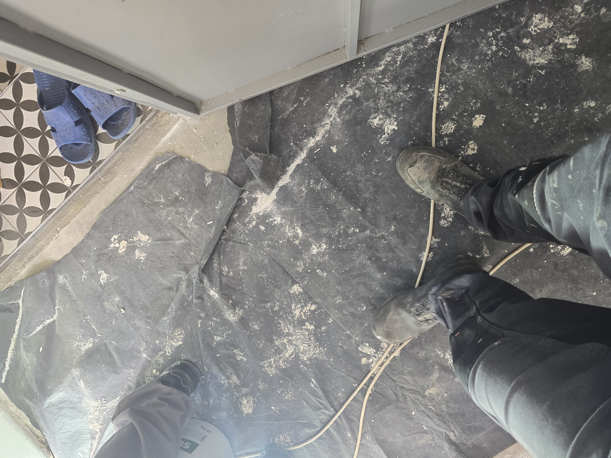 The image shows a person standing on a floor covered with a black plastic sheet. The floor appears to be dusty or covered with construction debris. The person is wearing dark-colored work boots and pants. A white electrical cord runs across the floor. To the left, there is a section of decorative tiled flooring. A pair of blue sandals are visible near the tile. The location is likely an indoor construction or renovation site. There are no cues to suggest a link to Gorno Aleksandrovo, Bulgaria.