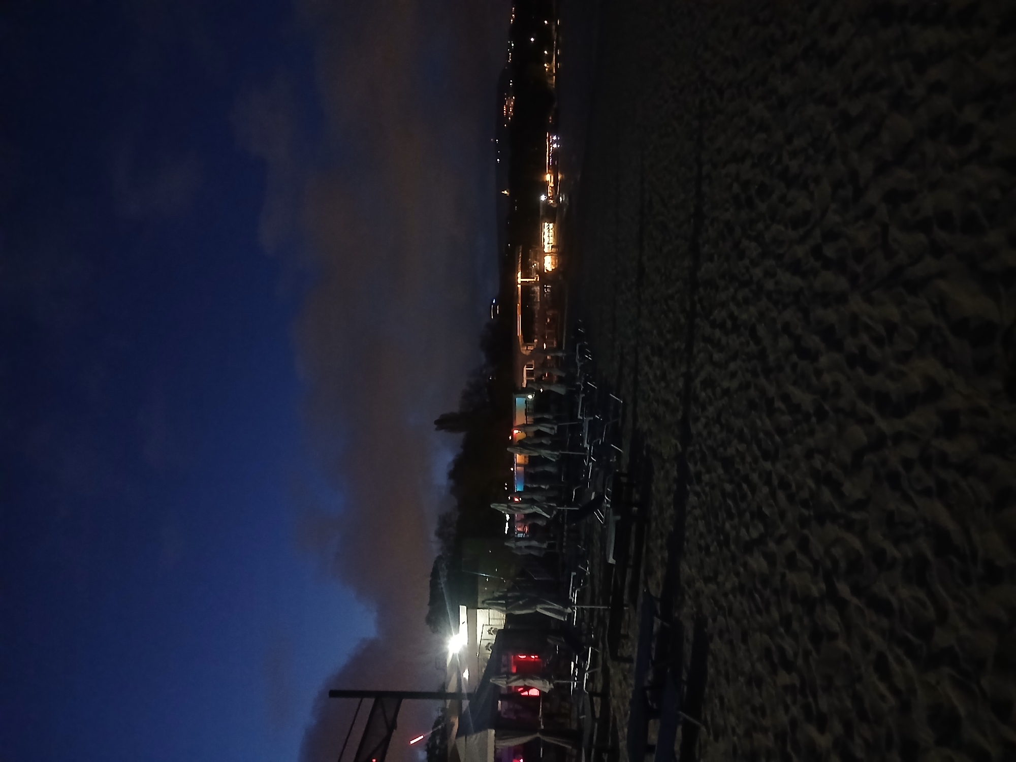 The image shows a nighttime scene on a beach in Varna, Bulgaria. In the foreground, there are rows of beach chairs, some with umbrellas, extending into the distance. The beach is made of sand and is dark. Further back, there is a building and lights. The background includes a view of Varna, with scattered lights visible in the distance. The sky is a dark blue color with clouds.