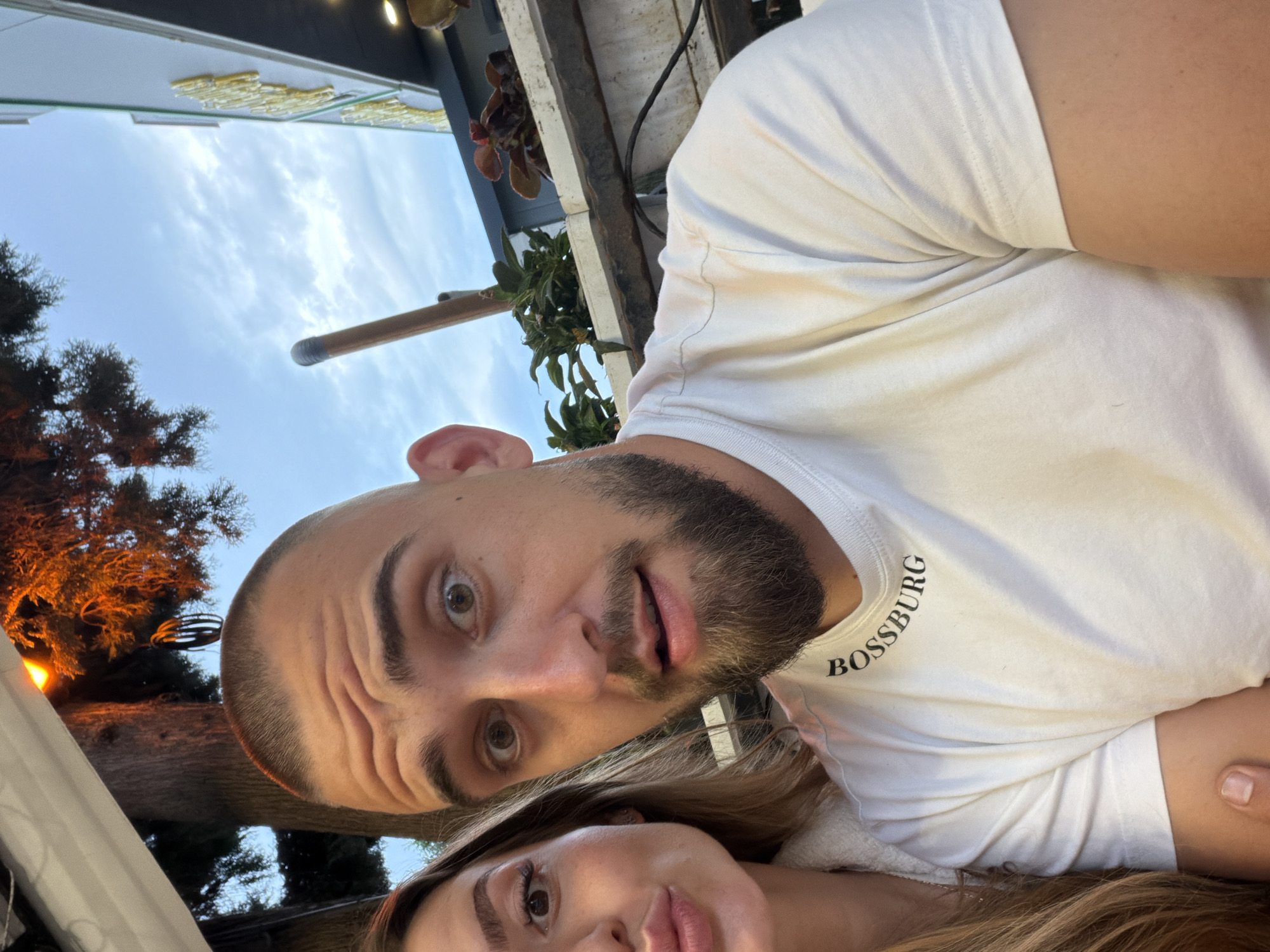 A selfie image shows a man and a woman posing close together. The man is bald with a full beard, wearing a white t-shirt with "BOSSBURG" printed on it. The woman has long, light-colored hair. The background includes sky, trees, and architectural details. The location might be Stara Zagora, Bulgaria.