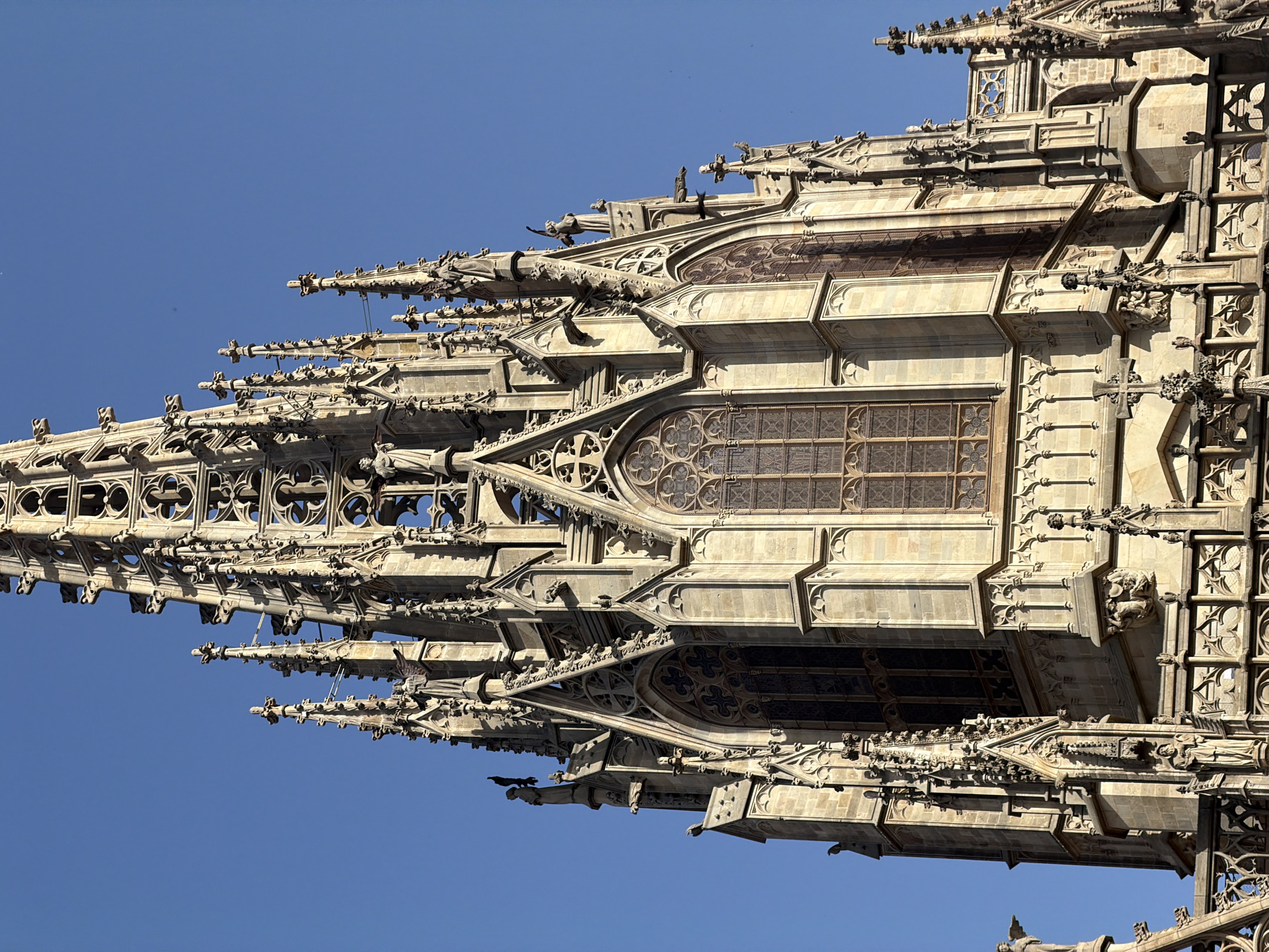 The image shows a close-up, angled view of a section of the Barcelona Cathedral's spires and upper facade against a clear blue sky. The architecture is Gothic, characterized by intricate stonework, pointed arches, gargoyles, and decorative tracery. Several statues are visible on the spires, along with ornate window structures and smaller pinnacles. The stone appears aged and weathered, casting shadows due to the sunlight. No people are visible in the image.