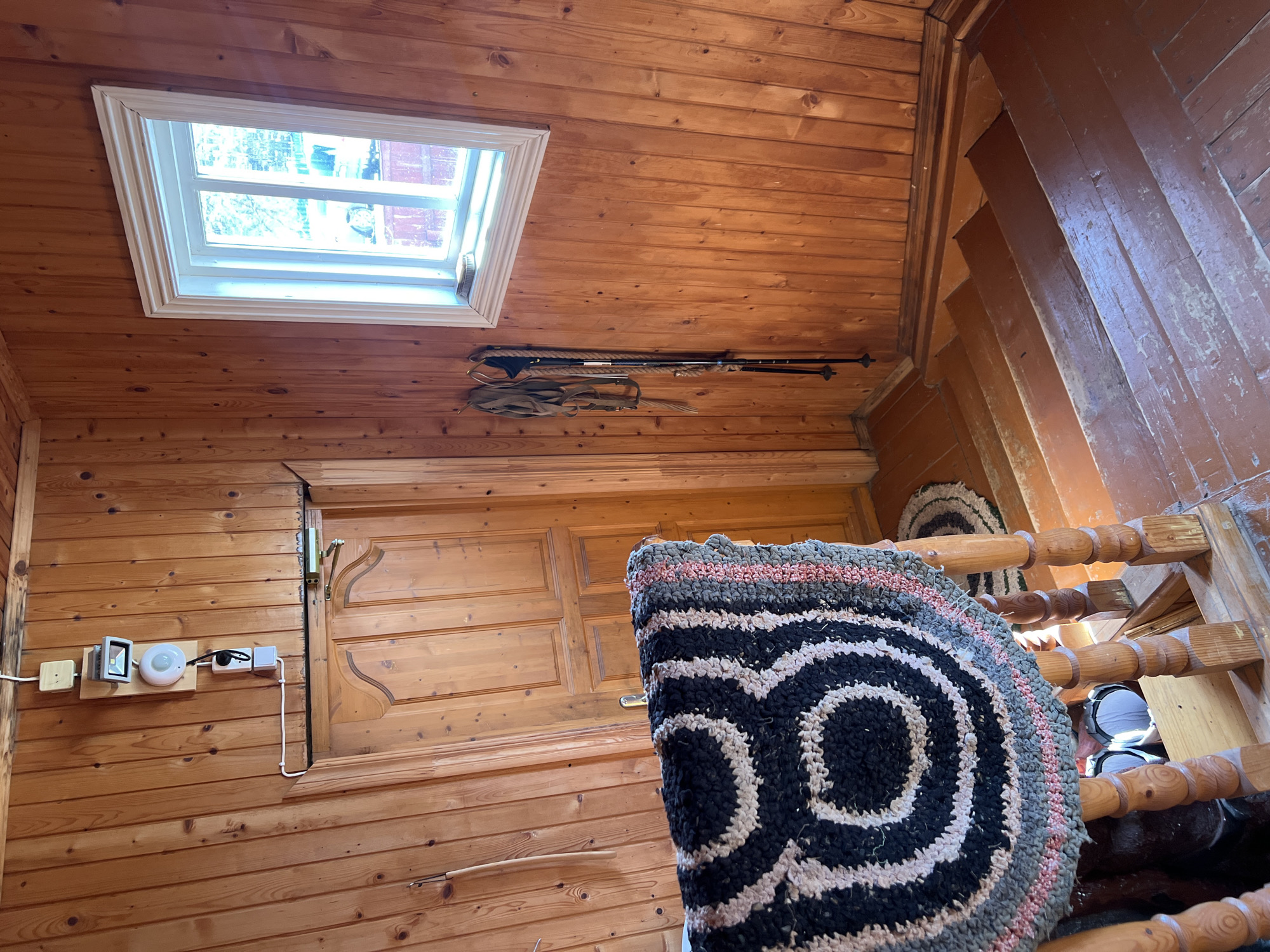 The image captures an interior view, likely within a residence, in Чернышово, Russia. The space is characterized by extensive use of wood, evidenced by the wooden walls, ceiling, and staircase. A rectangular window is situated on the ceiling, allowing natural light into the room. Adjacent to the window, on the ceiling, there's a pair of ski poles and associated straps. On the wall there's a light switch and a door with an ornate handle. A circular rug featuring a black and white concentric design is prominently displayed in the center, partially obscuring the view of a wooden staircase with a matching rug at the top. The stairs are painted brown.