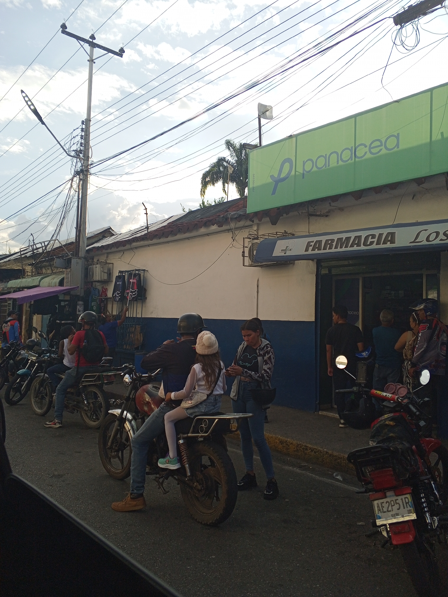 The image depicts a street scene in Tinaquillo, Venezuela. Several people are present, some riding motorcycles and others standing near a pharmacy named "Farmacia Los." A green sign with "Panacea" is visible above the pharmacy. Power lines are visible overhead, and several motorcycles are parked along the street. A woman appears to be looking at her phone. The image seems to have been taken from inside a vehicle. License plates are visible on at least one of the motorcycles.