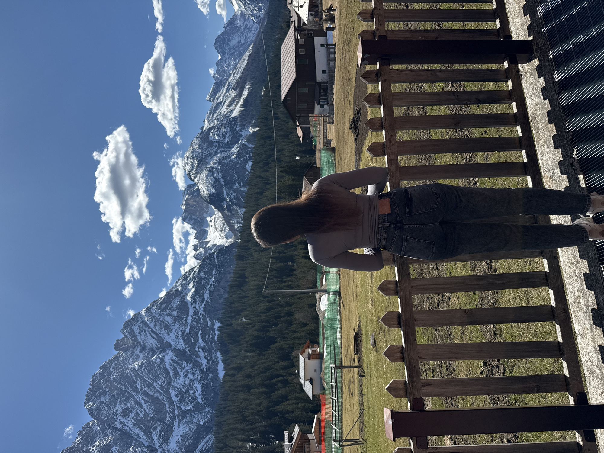 A medium, landscape-oriented shot captures a woman looking at a mountain view from a wooden platform or balcony. The scene is likely set in Sappada, Italy, given the mountainous terrain typical of the region.
The woman, positioned in the center of the frame, stands with her back to the viewer. She has long brown hair and wears a light gray long-sleeved shirt and dark gray jeans. Her hands seem to rest on the wooden fence or railing in front of her.
Behind the woman, a valley stretches out, featuring a mix of evergreen trees and grassy fields. Several buildings dot the landscape, including a house with a brown roof and white walls. The mountain range dominates the background, its rocky peaks partially covered with snow. The sky is bright blue with scattered white clouds.
In the foreground, the wooden fence runs horizontally across the frame, parallel to the woman's body. A section of the platform is visible, constructed from a metal grating. The lighting suggests a sunny day, casting shadows and creating highlights on the objects and surfaces.