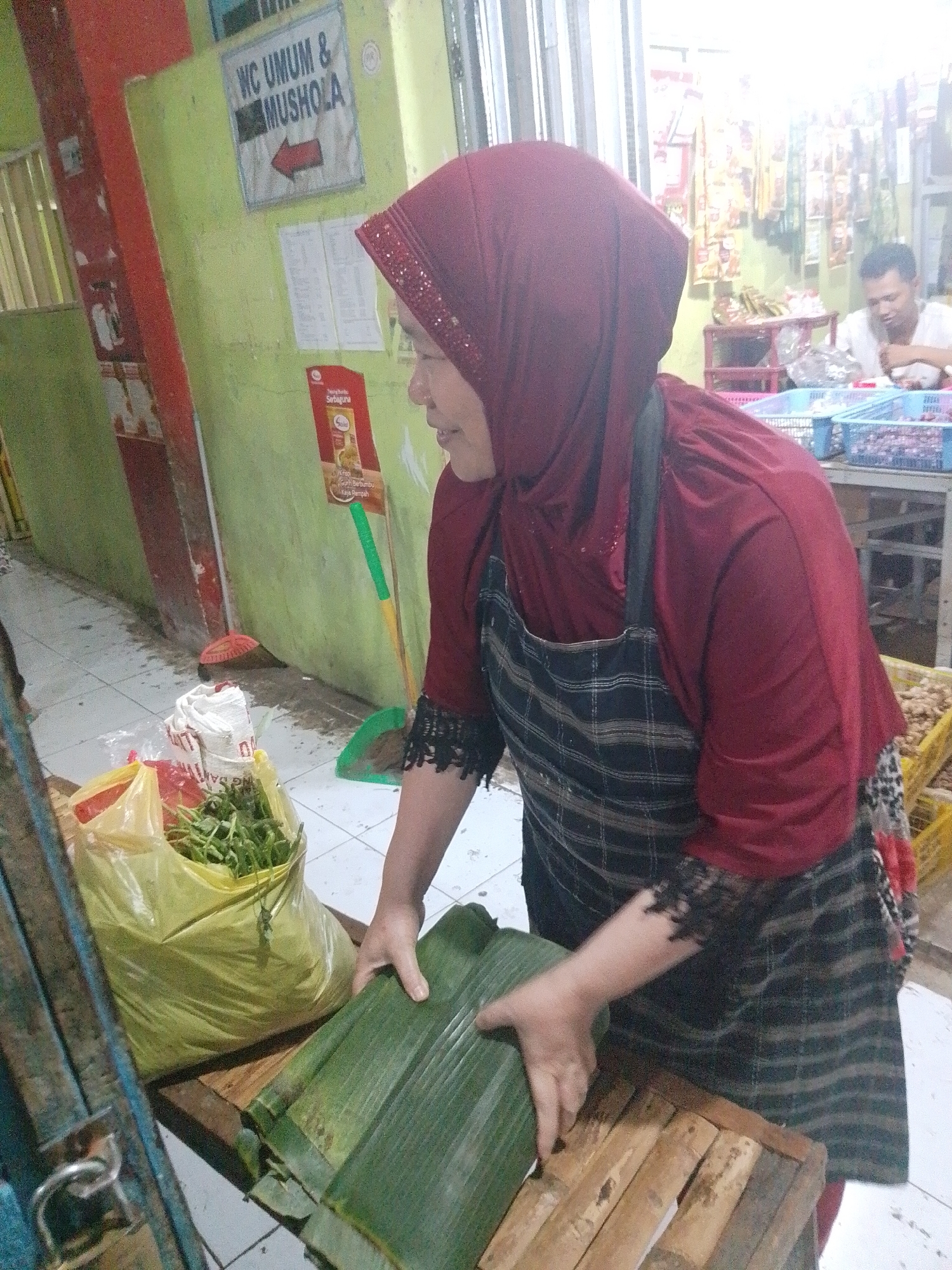An Indonesian woman, wearing a maroon hijab and an apron, is preparing food wrapped in banana leaves. She is standing in front of a wooden stall at what appears to be a market or food stall in Kecamatan Pati, Indonesia. To her left is a yellow plastic bag filled with green vegetables. Behind her, a sign reads "WC UMUM & MUSHOLA" with an arrow pointing left. In the background, a man in a white shirt is visible, along with shelves stocked with various goods. The environment suggests an outdoor or semi-outdoor market setting with a tiled floor. The lighting is bright, possibly from overhead lights.