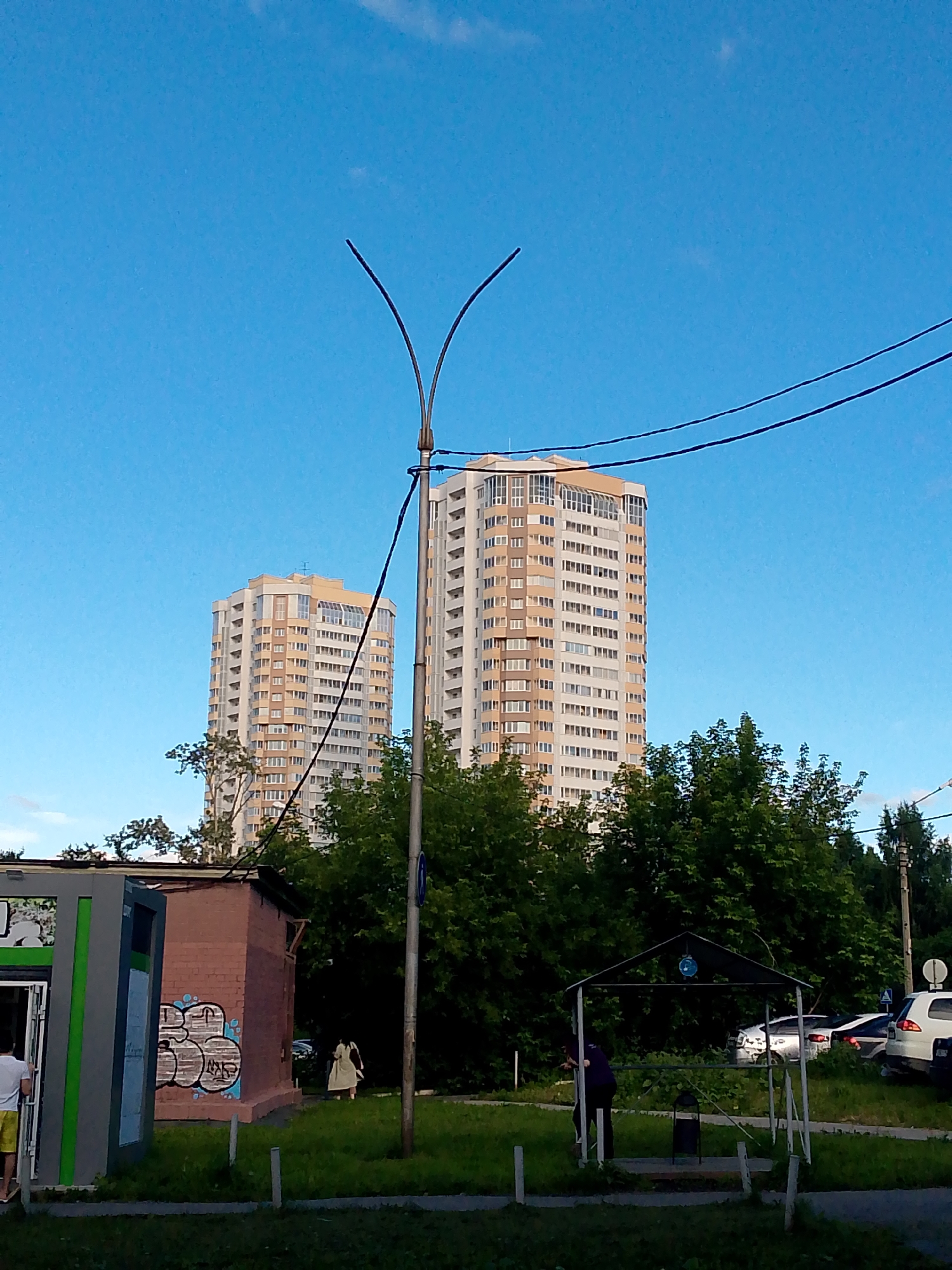 A clear blue sky is visible above a cityscape. In the mid-ground, two tall, multi-story buildings with beige and white facades stand behind a dense line of green trees. The building on the right is taller than the one on the left. In the foreground, a man in a white t-shirt and yellow shorts is seen entering a grey and green structure on the left. Adjacent to this structure is a small brick building with graffiti on its side. Further to the right, a person in a light-colored dress walks away from the camera. Behind the trees and to the right, a covered structure with a dark roof and white supports is visible, with a trash can underneath it. Several cars are parked in the background. A street lamp with two curved arms is in the foreground, with black wires extending from it across the frame.