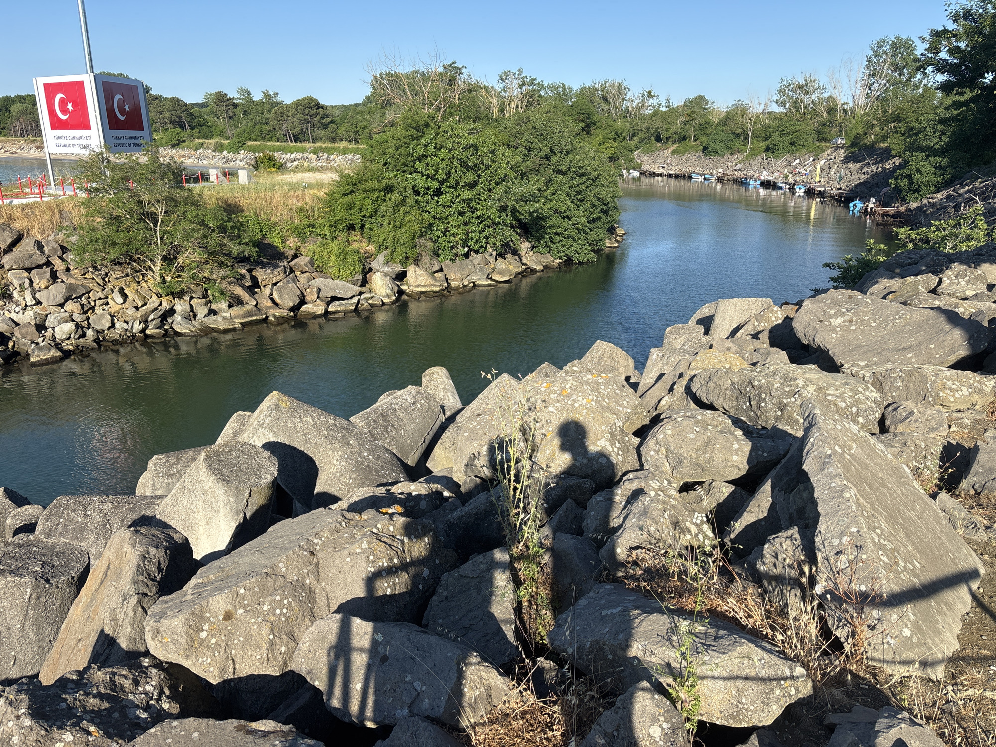 The image depicts a scene in Rezovo, Bulgaria, likely near the border with Turkey. In the foreground, a pile of large, weathered gray rocks forms a breakwater or pier. The rocks are irregularly shaped and some are covered in lichen. In the mid-ground, a body of water, possibly a river or inlet, flows between rocky banks. On the left bank, a sign with the Turkish flag and text "TÜRKİYE CUMHURİYETİ REPUBLIC OF TURKEY" is visible, indicating proximity to the Turkish border. Further into the water, small boats are moored along the right bank, which is lined with trees and vegetation. In the background, more trees and vegetation cover the landscape under a clear blue sky. Shadows of people and what appears to be a fence are cast across the rocks in the foreground.