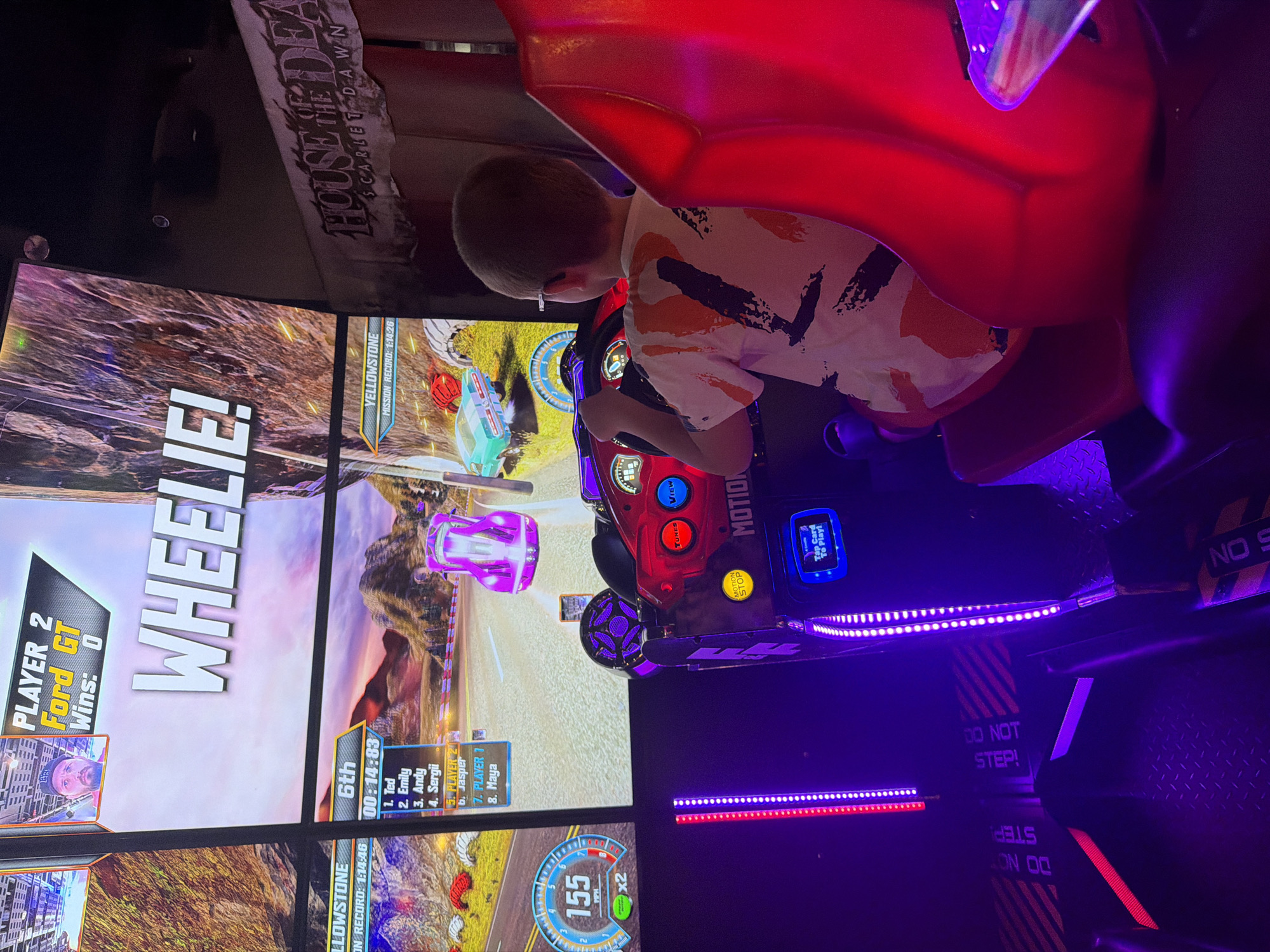 A young boy with light brown hair and glasses is playing a racing arcade game. He is seated in a red plastic racing seat and gripping a black steering wheel. The game screen shows a car racing on a road with a mountainous landscape in the background. The game displays "WHEELIE!" prominently at the top. The arcade cabinet has various buttons and a small screen at the bottom. The area is dimly lit, with purple and red LED lights illuminating the surroundings, including the floor which has "DO NOT STEP" warnings printed on it. In the background, a banner for "HOUSE OF THE DEAD SCARLET DAWN" is visible. The scene is likely in an arcade or entertainment venue in Prague, Czechia.