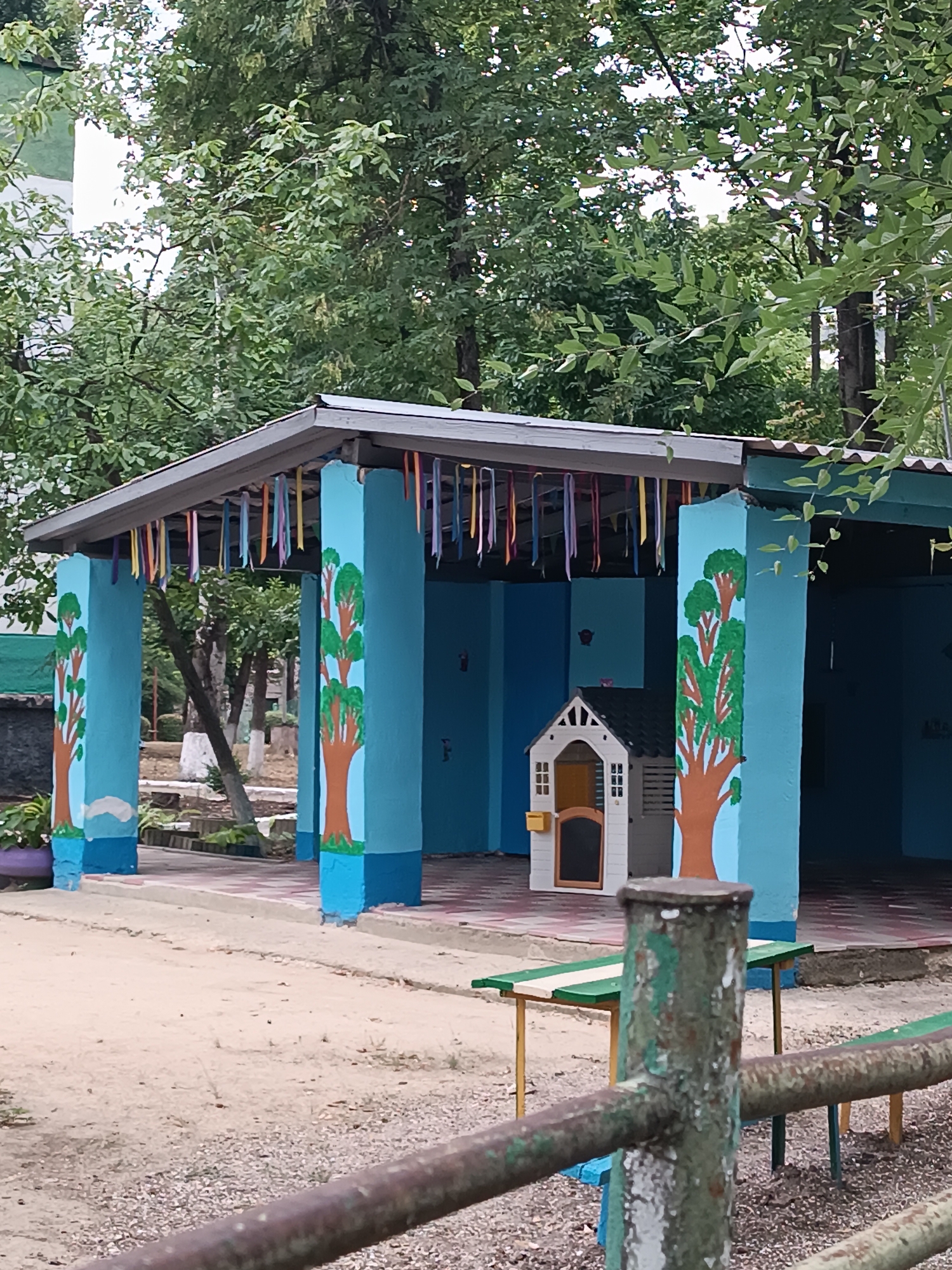 The image depicts an outdoor play area, likely for children, under a covered structure. The structure has a grey roof supported by blue pillars painted with tree motifs. Colorful ribbons are hanging from the edge of the roof. In the background, there's a small white playhouse with a yellow mailbox. A simple table with green and white stripes and a bench are visible in front of the structure. The ground is a mixture of sand and gravel. In the foreground, a rusty metal fence runs horizontally across the frame. The scene is surrounded by trees with green foliage, suggesting a temperate climate. The architectural style and the presence of a play area imply a location such as a kindergarten, school yard, or public park within a residential area. Given the request to specify the location in Chișinău, Moldova, it is plausible that this image was taken at a facility or public space in the city. However, without further information or distinguishing landmarks, it is impossible to pinpoint the exact location within Chișinău. The overall impression is one of a simple, functional outdoor recreational space.