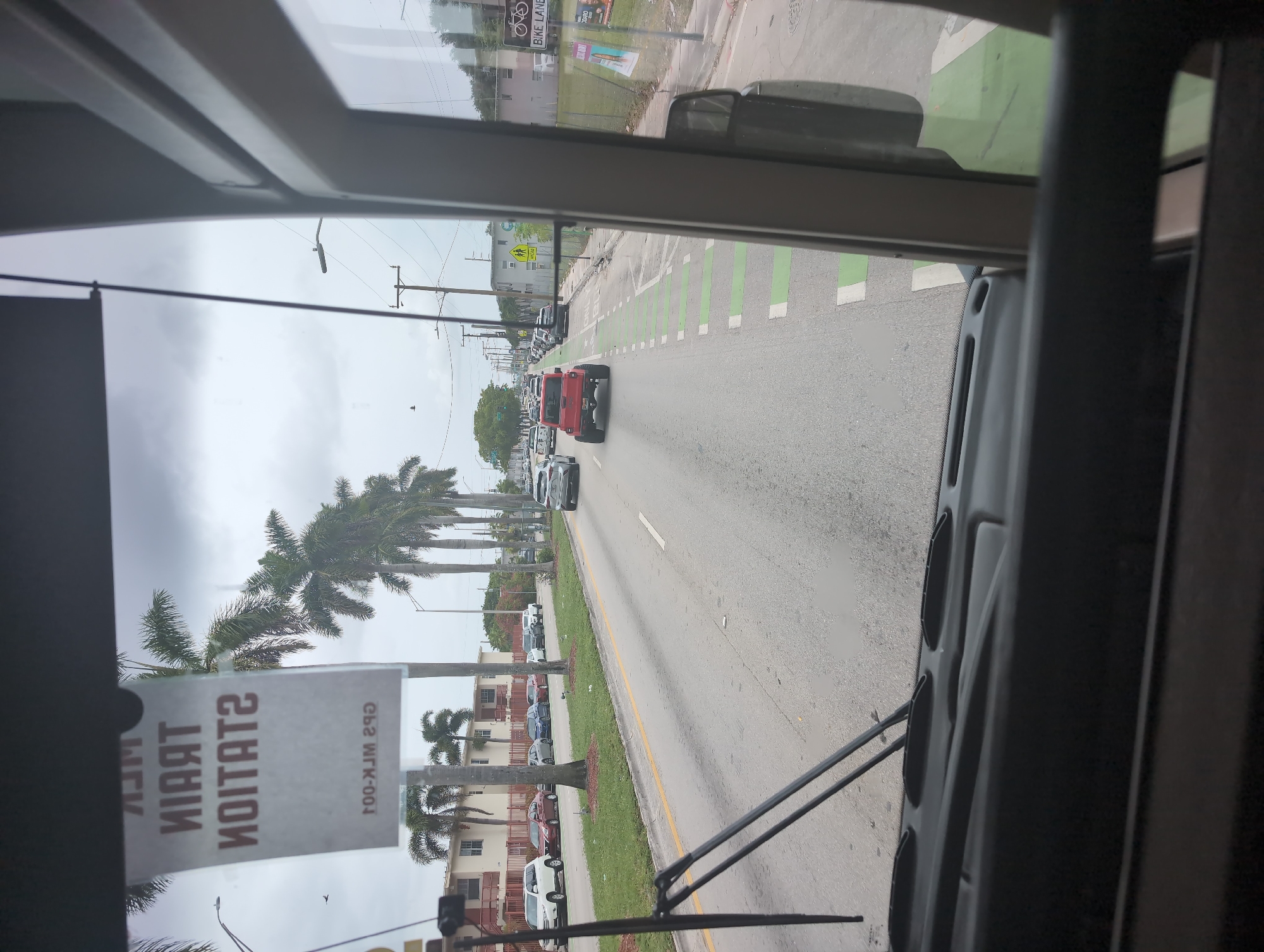 The image shows a view from inside a bus looking out the front windshield and side window. The bus is driving on a street in Opa-locka, Florida. Palm trees line the street, and there are several cars and buildings visible. A sign hangs from the windshield that reads "TRAIN STATION" upside down. A red truck is driving in front of the bus, and there is a bike lane marked with green paint on the right side of the road. The sky is cloudy.