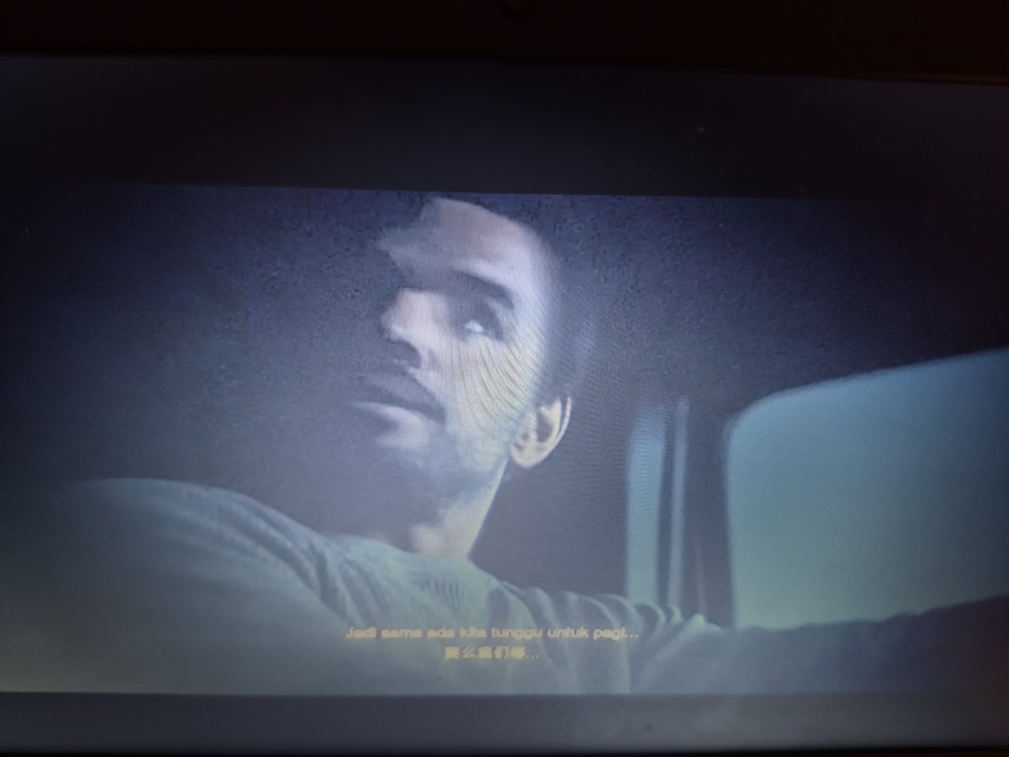 The image is a close-up of a television screen displaying a scene from a movie or show. The screen shows a close-up of a person's face from the shoulders up. The person is looking upwards and to the left. The lighting is dim, with a light source appearing to be from the right. The person is wearing a light-colored shirt. Subtitles are visible at the bottom of the screen. The top portion of the image shows the dark bezel of the television screen. There are no discernible elements in the image that would indicate a location within the city of Sandwell, United Kingdom.