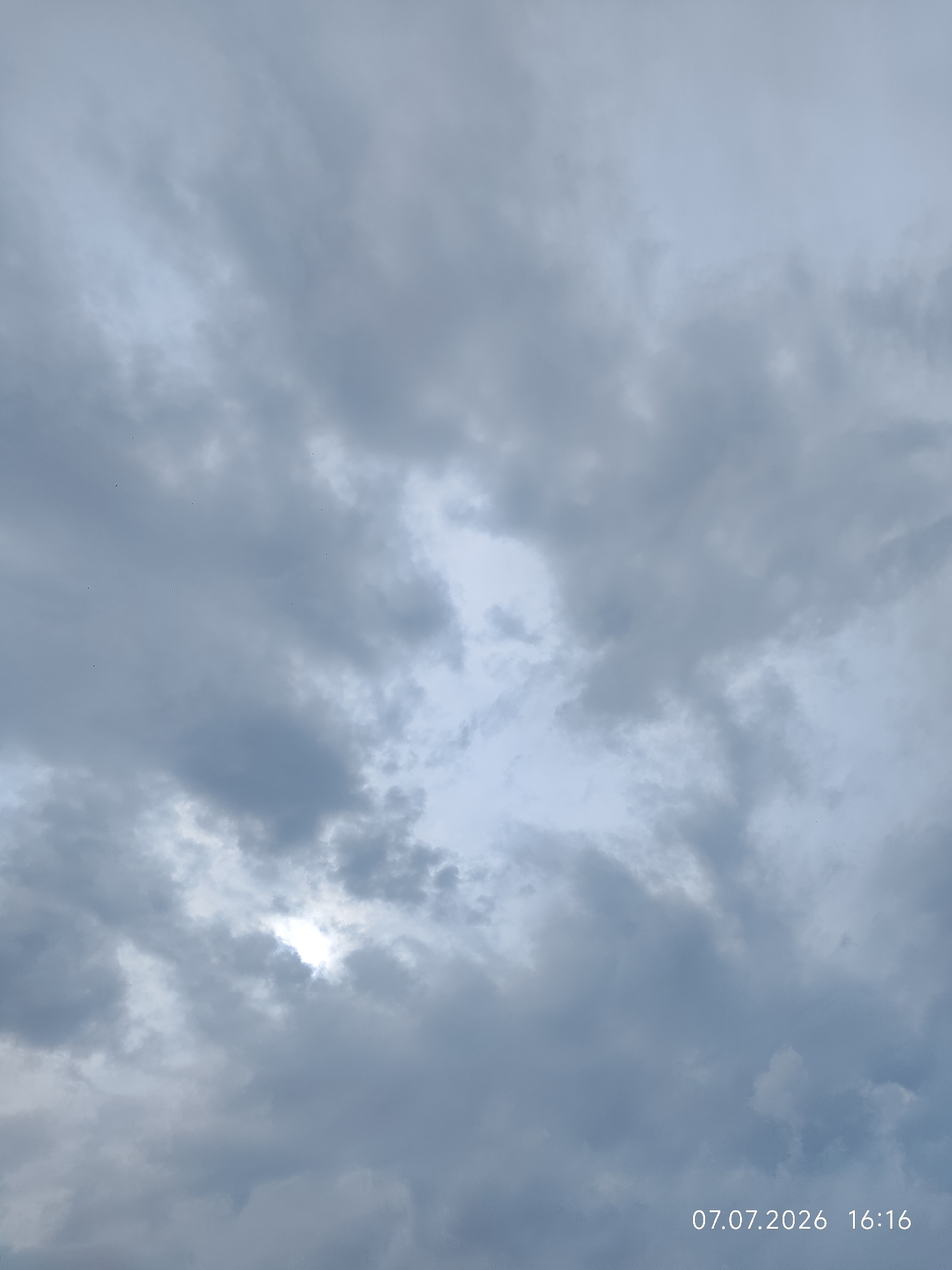 The image displays a cloudy sky over Konovalovo, Russia, on July 7, 2026, at 16:16. The sky is filled with various shades of gray clouds, with patches of lighter sky visible through some breaks. The sun is partially obscured by the clouds, creating a soft illumination. No people, subjects, or objects are clearly identifiable in the scene. The environment is solely characterized by the atmospheric conditions.