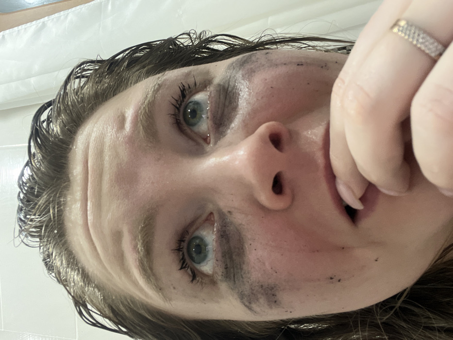 An upward angle close-up shows a woman's face with smudged black marks around her eyes and cheeks. Her hair is wet and slicked back. Her expression is somewhat distressed, with wide eyes. A hand with a ring on one finger is near her mouth. A white surface, possibly a shower or tile wall, is in the background. The lighting is bright and harsh.