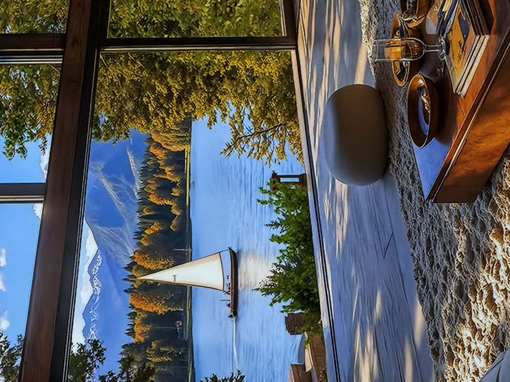 The image displays an interior setting with a large window offering a panoramic view of a natural landscape, rotated 90 degrees counter-clockwise. When correctly oriented, the left side of the frame is dominated by the window, framed in dark wood.
Through the window, a picturesque scene unfolds. A wide, calm body of water, likely a lake or river, occupies the middle ground, reflecting the clear blue sky. Its shores are lined with dense forests, predominantly showcasing vibrant autumn foliage in shades of yellow and orange, intermingled with dark green conifers. In the distant background, majestic, snow-capped mountains rise under a bright blue sky with scattered white clouds. A sailboat with a distinctive white, triangular sail, on which a small number "1" is visible, glides across the water. Green tree branches from foreground trees are seen close to the window, partially framing the view.
The interior, on the right side of the window, features a light-colored floor with prominent parallel shadows cast by sunlight, indicating a bright daytime setting. Part of the floor is covered by a textured, light-colored rug with an intricate pattern. On this rug, a smooth, dark grey, oval-shaped object is placed. Further right, a dark wooden tray holds a clear glass containing an amber liquid (possibly wine or a spirit), a small brown bowl, and a stack of rectangular items, possibly books or decorative boxes. No people are visible in the scene. The overall atmosphere conveys tranquility and appreciation for natural beauty. The provided location context of Sofiivska Borshchahivka, Ukraine, is not directly discernible from the image's visual cues.
