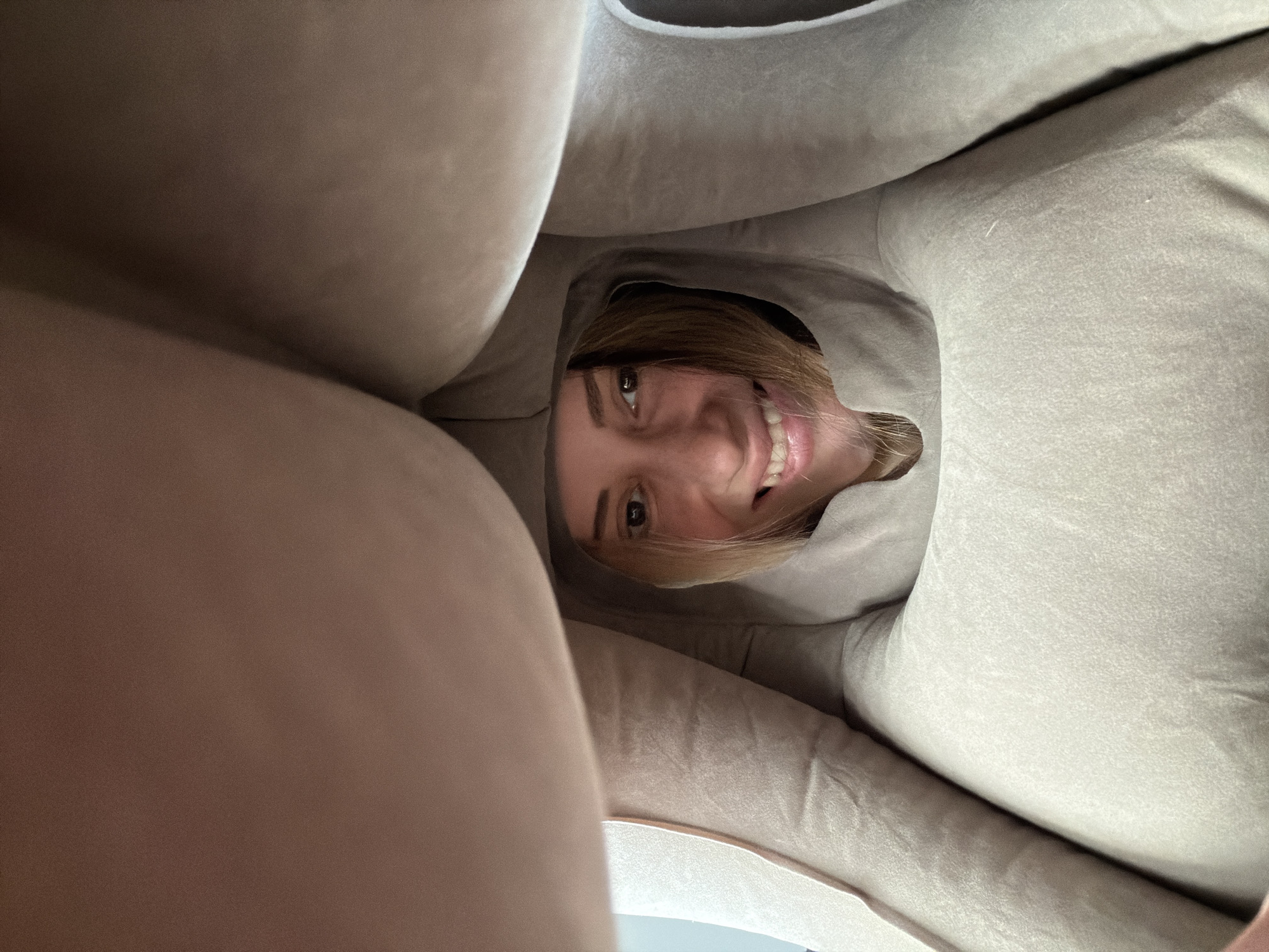A young, fair-skinned woman with brown eyes and blonde hair smiles directly at the camera from within a soft, beige or light brown fabric enclosure. The fabric is bunched around her face, creating a tunnel-like perspective. The image is taken from a low angle looking upward at the woman. The environment appears to be indoors. There is no information to suggest the image was taken in совхоза "Воронежский", Russia.