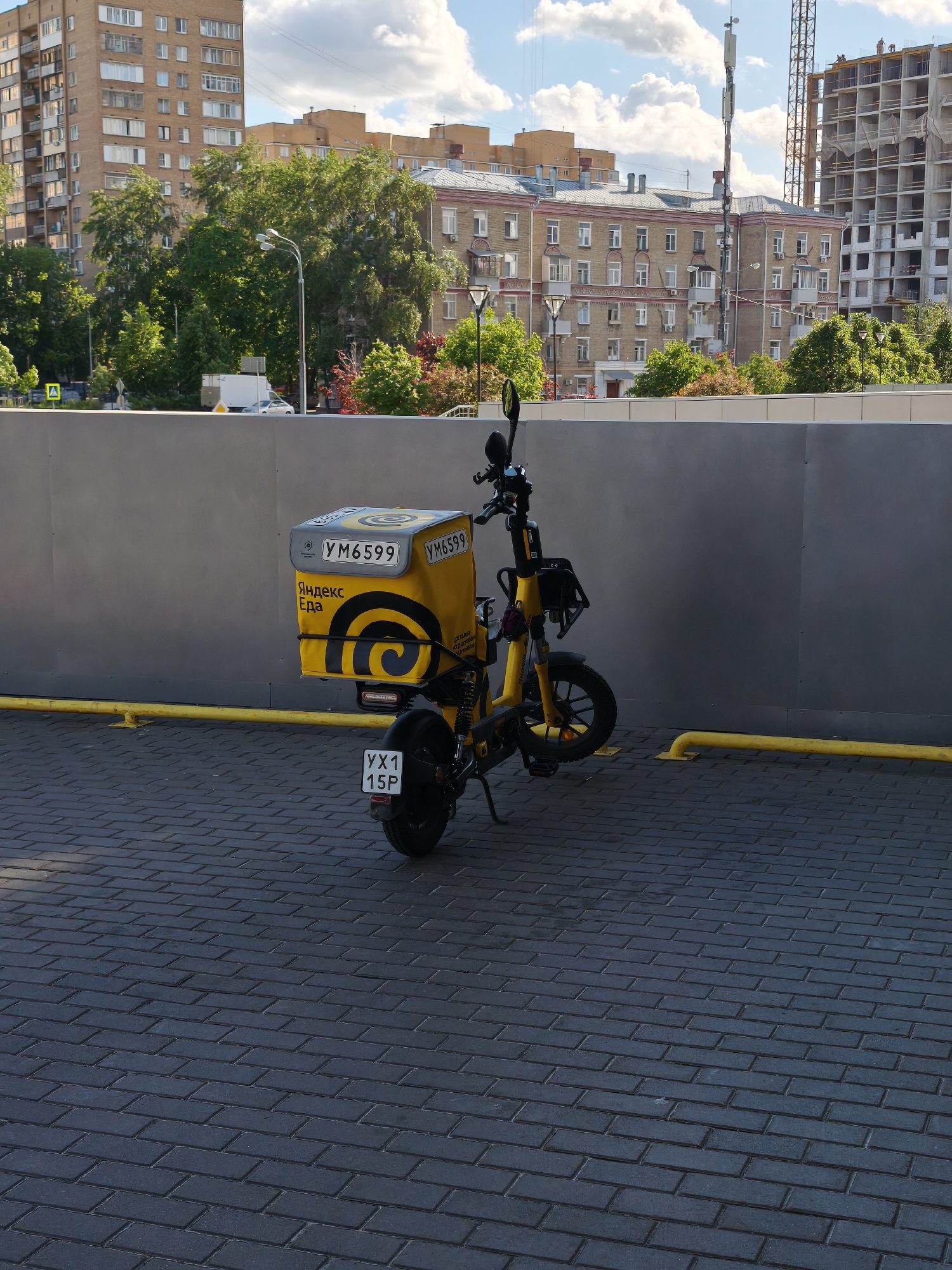 The image shows a yellow Yandex.Eda delivery scooter parked on a brick-paved surface against a grey concrete wall. The scooter has a box with the "YM6599" license plate on it, along with the Yandex.Eda logo. The scooter has the license plate "YX1 15P".
In the background, there are several multi-story apartment buildings, some trees, a streetlight, a communication tower, and a construction site with a crane.
Based on the architecture and the presence of the Yandex.Eda scooter, the location is likely in Moscow, Russia.