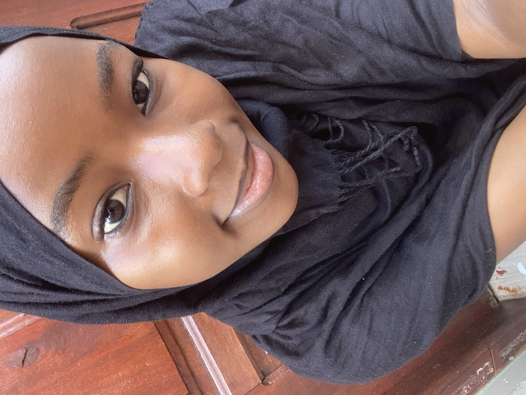 A close-up, slightly high-angle portrait features an individual, appearing to be a young female, centered in the frame. The subject has a dark skin tone and is wearing a black headscarf (hijab) and a black garment, with visible fringes on the chest area. Her dark eyes are open and directed forward, with well-defined dark eyebrows. A subtle closed-mouth smile is present. The skin on her face exhibits a luminous quality, particularly across the forehead and cheeks. The background consists of a reddish-brown wooden surface, likely a door or paneled wall, displaying vertical lines, wood grain, and minor surface imperfections. A lighter, possibly concrete or tiled surface with reddish-orange stains is visible at the lower left edge. While the image is presented in the context of Dar es-Salaam, Tanzania, the specific location within the city cannot be determined from the visual information provided.