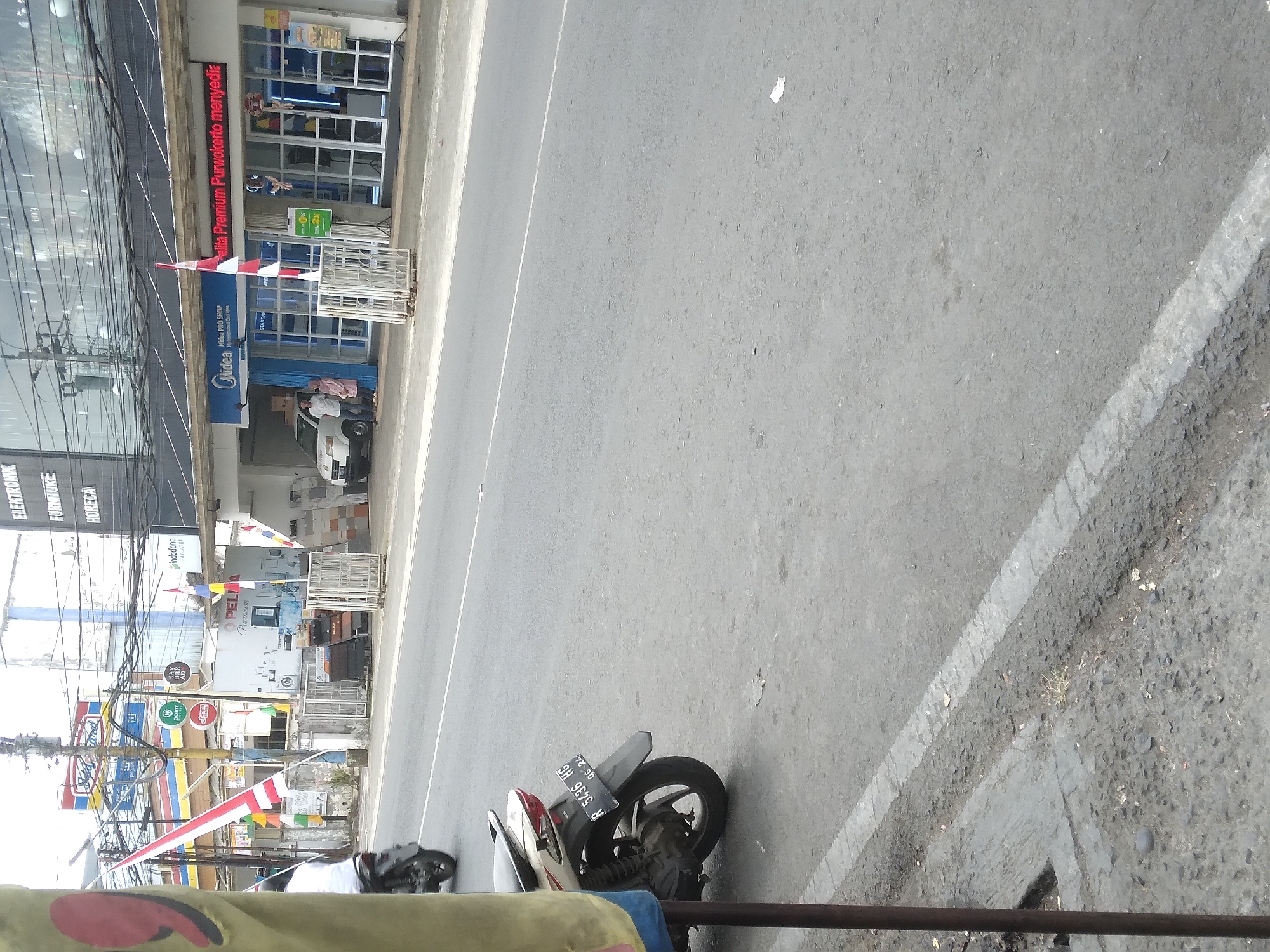 A medium shot captures a street scene in Berkoh, Indonesia, dominated by a grey asphalt road and a concrete sidewalk on the left. On the right side of the road, a row of buildings forms the backdrop, featuring storefronts with large glass windows. One prominent establishment is labeled "Midea PRO SHOP," displaying the Midea logo and text in Indonesian. Adjacent to it, another storefront is illuminated with red LED text that reads "Elita Premium Purwokerto menyediakan."
Several Indonesian flags, red and white, are visible, one attached to a pole in front of the Midea shop and others near the buildings. A white van with a couple of people standing near it is parked in front of the Midea shop. Two white metal barriers are positioned on the sidewalk.
In the foreground, a white and beige scooter is parked on the sidewalk, its license plate reading "R 5436 HG" and a date stamp "06-24." To the far left, a portion of a yellow and red striped object, possibly a banner or flag, is partially visible. Power lines crisscross overhead, and various signs and logos, including "Indomaret," "Point," and "J.Co," are discernible on the buildings. The overall atmosphere suggests a commercial area.