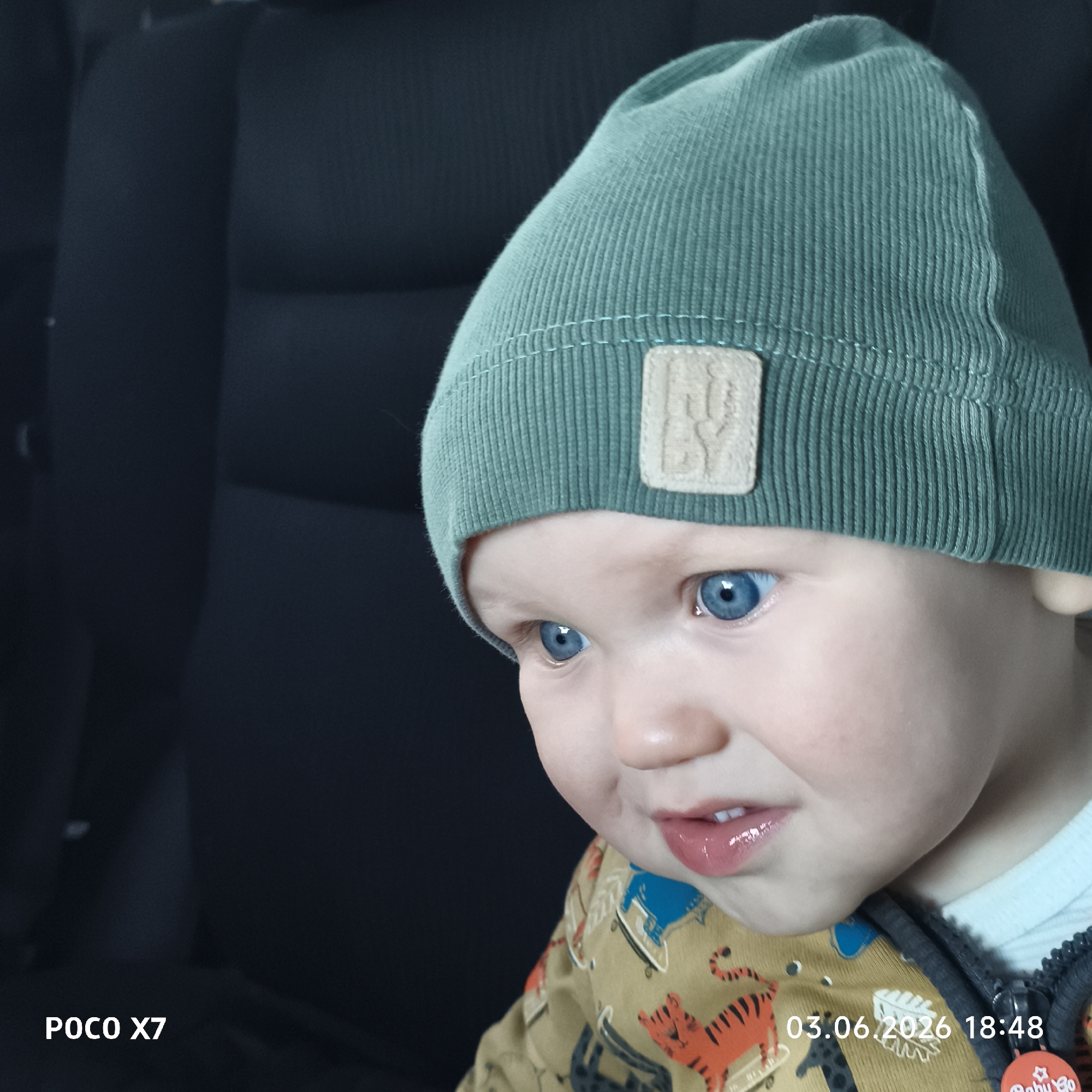 A close-up portrait features a young child, likely a toddler, with bright blue eyes and fair skin. The child is wearing a teal-green ribbed knit beanie hat with a light brown square patch on the front, featuring an indistinct, embossed logo. They are also dressed in a beige or light brown top adorned with cartoon-like animal patterns, including an orange tiger.
The child's mouth is slightly open, revealing some small teeth, and their expression is somewhat ambiguous, possibly indicating slight curiosity, grumbling, or a fleeting moment of contemplation. The background is dark and out of focus, suggesting the interior of a vehicle, likely showing car seats or upholstery.
Visible text in the bottom left corner reads "POCO X7", indicating the camera model. A timestamp "03.06.2026 18:48" is displayed in the bottom right corner. A small, red, circular tag with partially visible text, possibly "Baby Go", is attached to the child's clothing on the far bottom right edge. The image captures a candid moment of the child in what appears to be an indoor or vehicle setting during the evening.